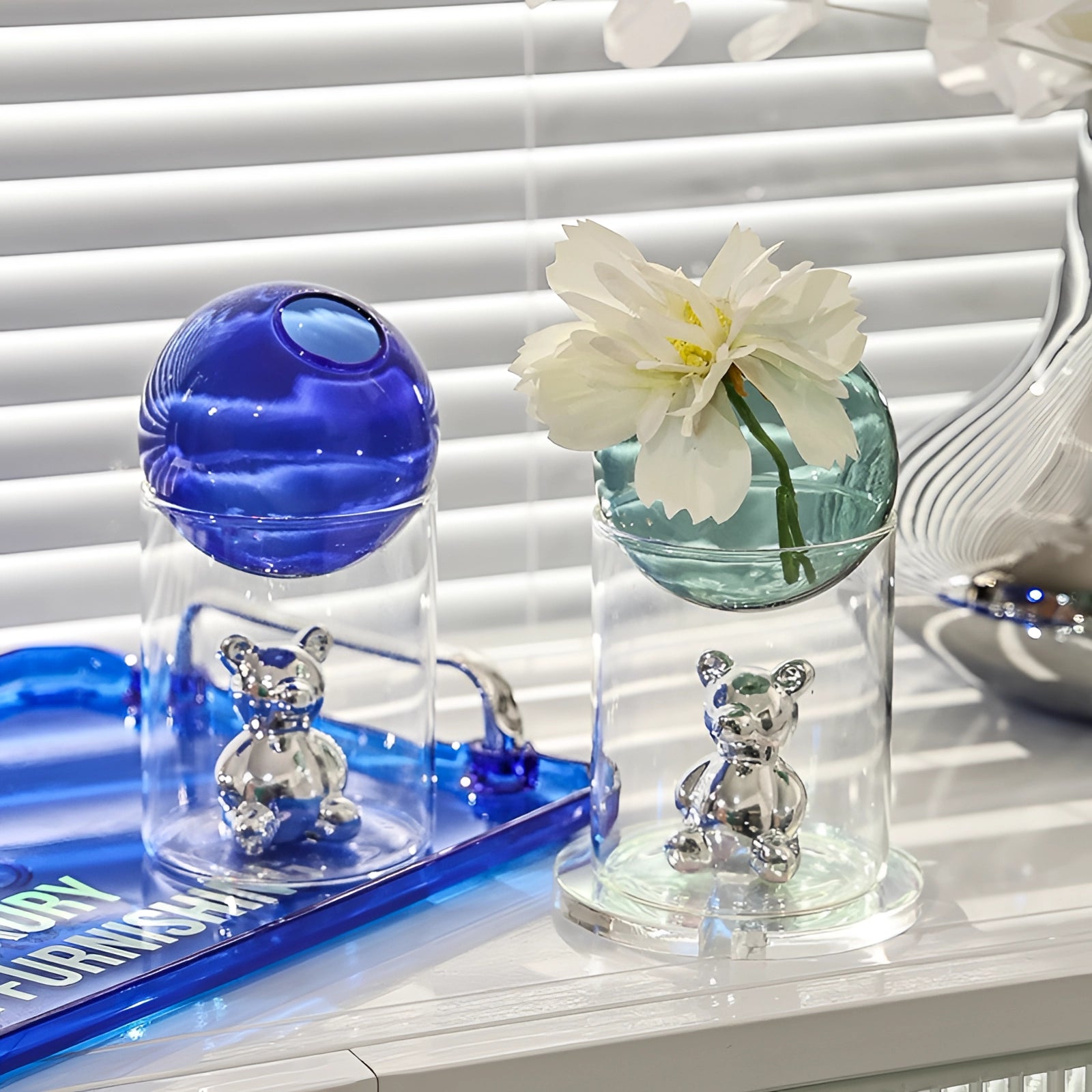 A luxurious amber glass sphere and gold bear decor piece displayed on a shelf, surrounded by vibrant colored elements including blue and purple drinkware and serveware, with accents of yellow and aqua, creating a sophisticated and elegant setting.