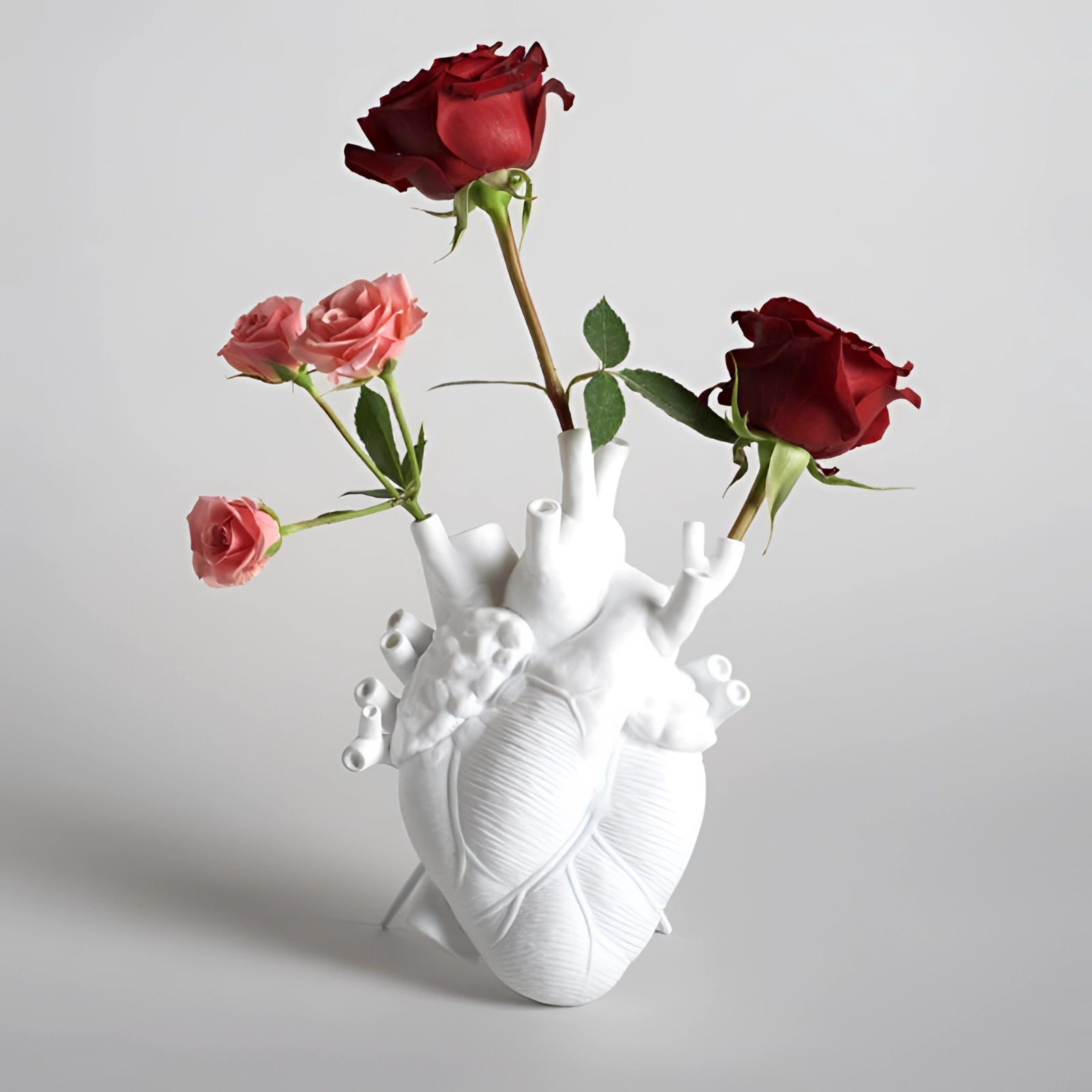 
A large, white anatomical heart vase functioning as a romantic ceramic floral sculpture, creatively arranged with intricately detailed artificial flowers resembling hybrid tea roses and other petals, forming a stunning bouquet.