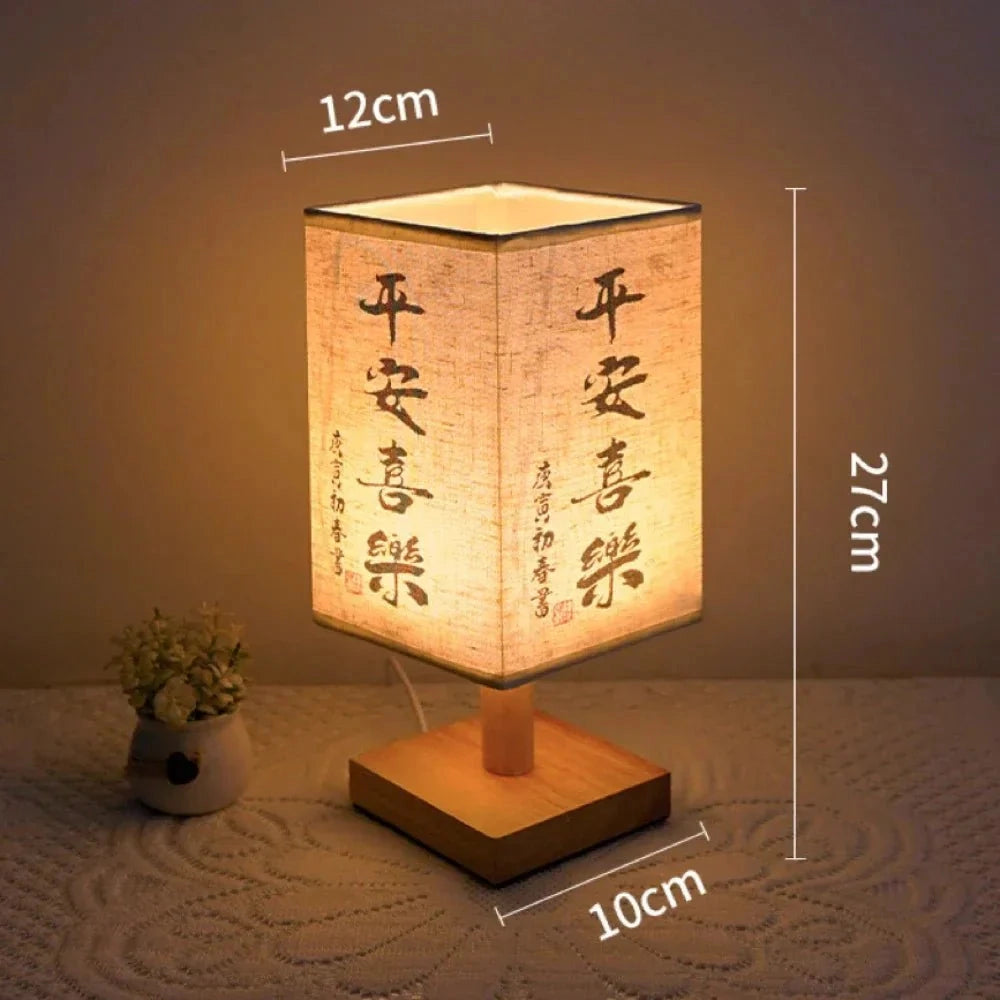 Asian calligraphy lamp with a wooden base and a compact design measuring 27cm, featuring a unique calligraphy design. Comes with a USB connector and serves as an elegant nightlight. The lamp is placed on a rectangular wooden base and surrounded by decorative elements like houseplants and flowerpots.