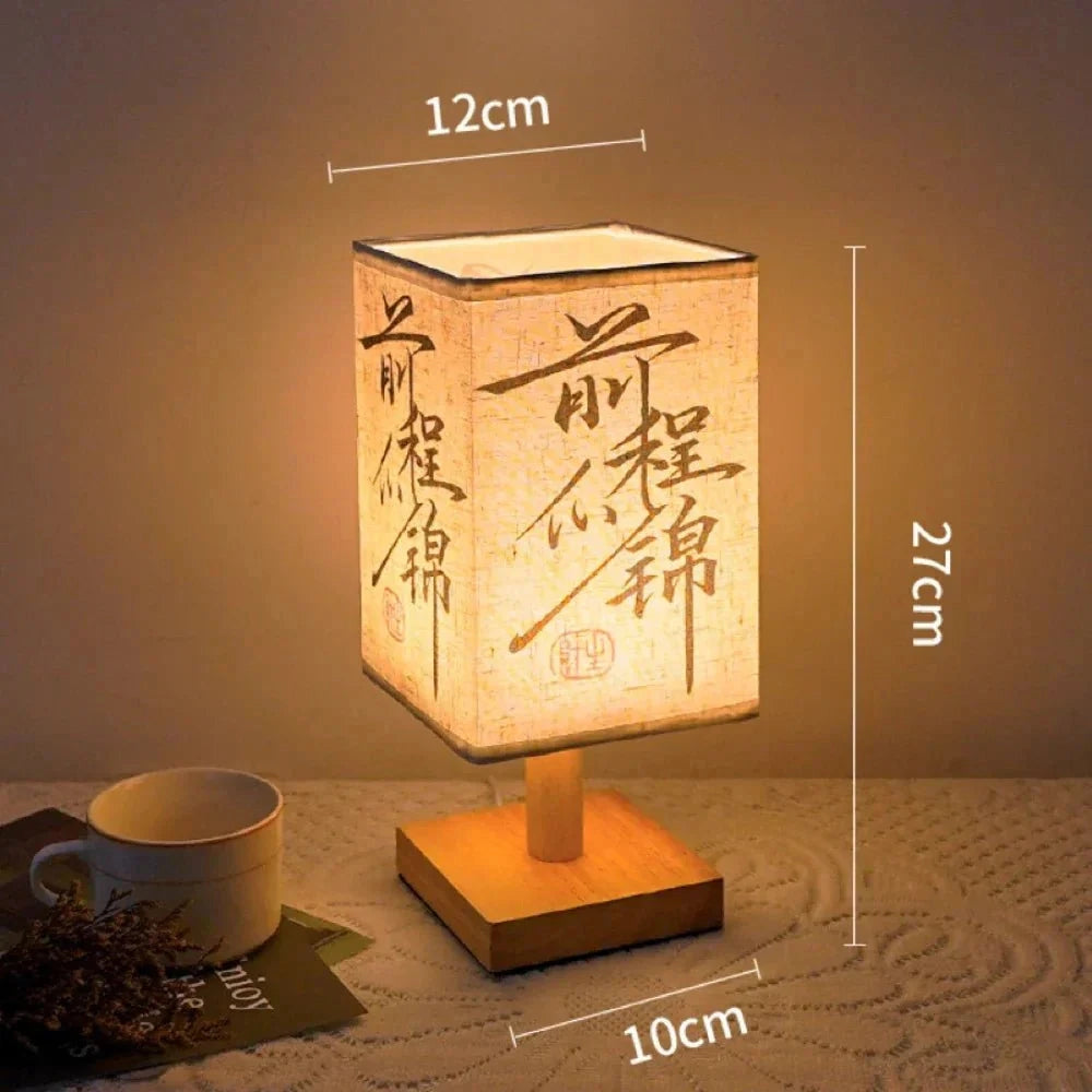 Asian calligraphy lamp with wooden base and compact design, measuring 27cm, featuring intricate handwriting art on its surface. The lamp is shown in a dimly lit room, emphasizing its warm glow. It has a USB connector, suitable as a nightlight or decorative piece on a table.