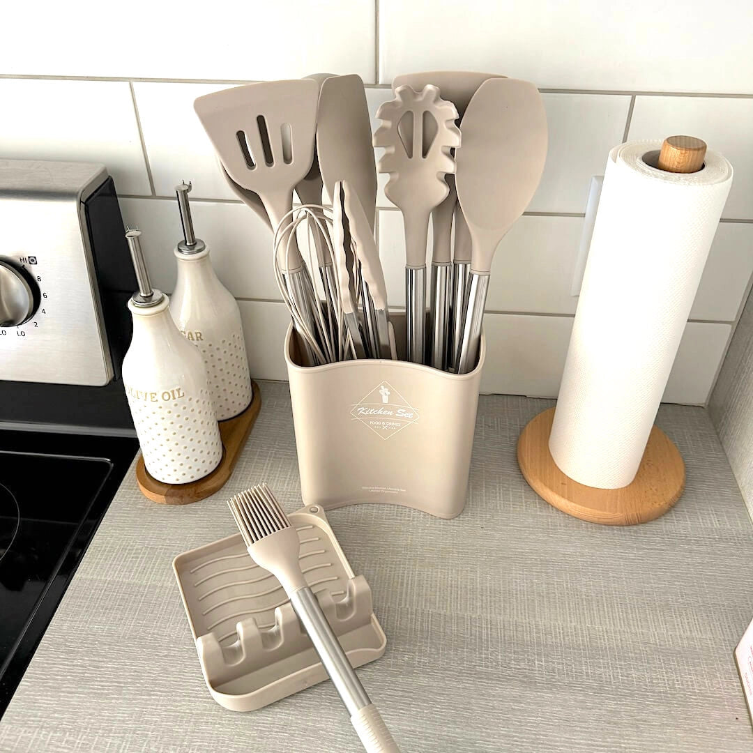 Beige silicone kitchen utensil set consisting of 12 pieces and a matching bucket, displayed on a wooden floor. The utensils are designed to be heat resistant and non-stick, suitable for various cooking needs.