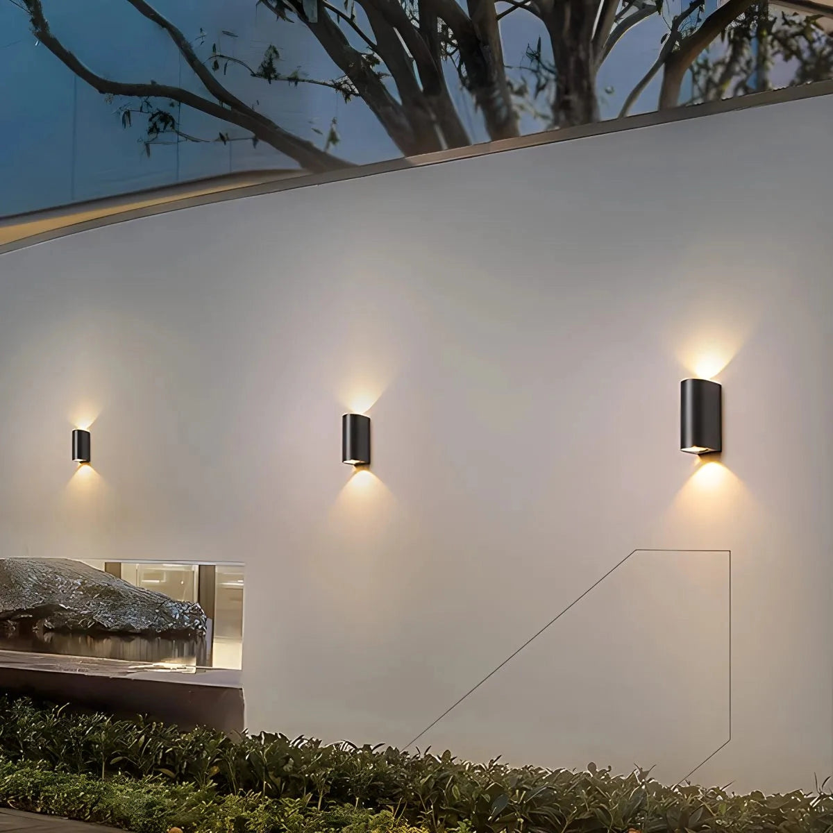 Modern black cylindrical LED outdoor wall lamp mounted on an exterior wall, illuminating the surrounding area with a warm yellow light, highlighting nearby plants and architectural features.