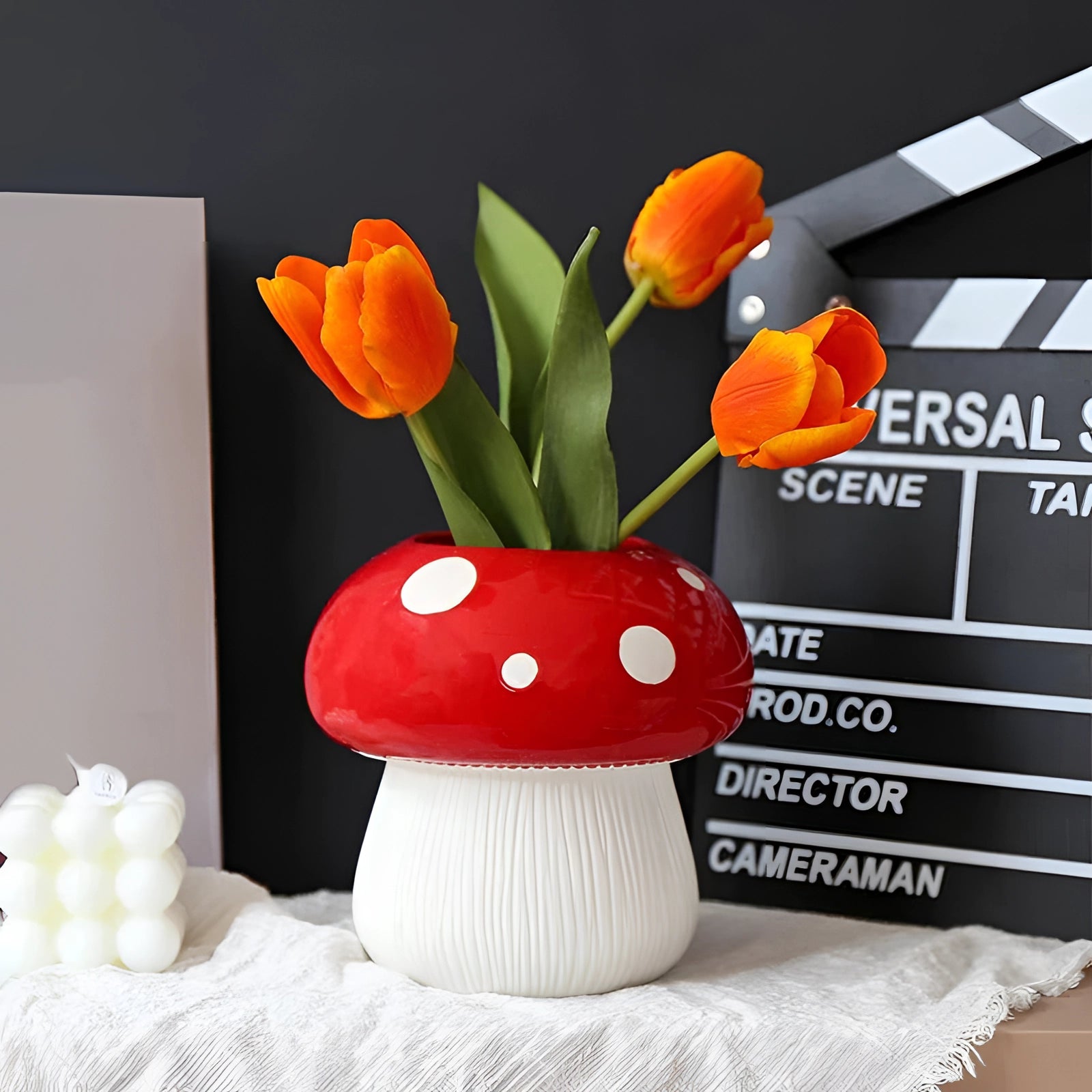 Black and white polka dot ceramic mushroom table lamp placed on a wooden table, surrounded by decorative objects including a vase with orange flowers and potted houseplants.