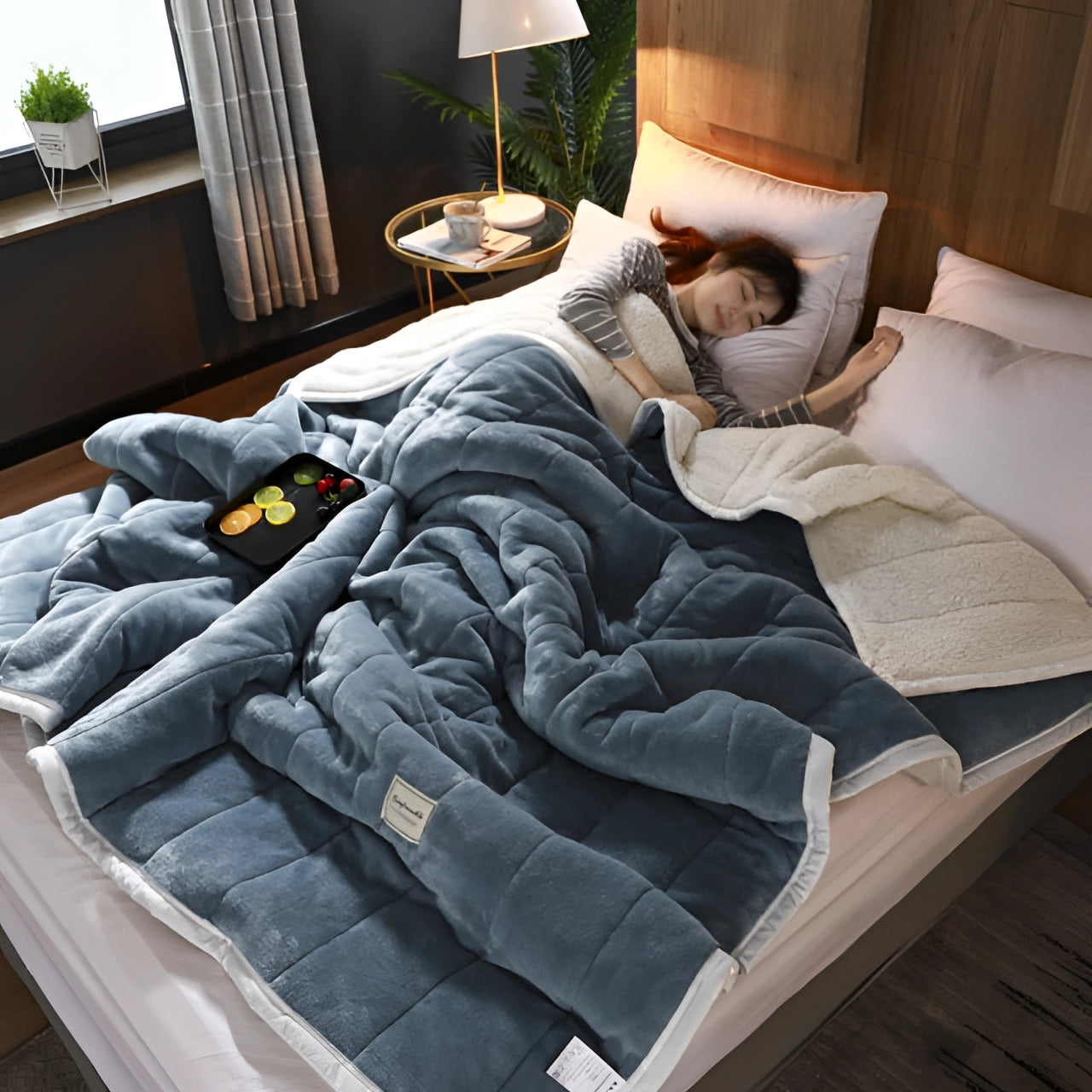 Blue plush weighted blanket in queen size draped over a bed in a cozy, well-lit room with wooden flooring, houseplants, and light-colored curtains, creating a comfortable and inviting interior design.