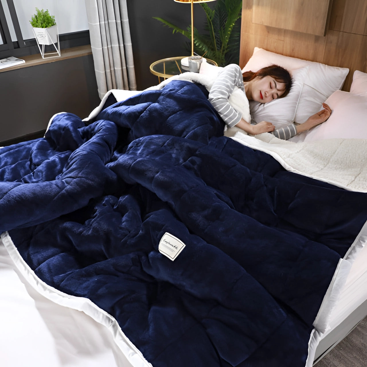 Blue plush weighted blanket draped over a neatly made queen-sized bed in a cozy bedroom setting, featuring marine blue color tones and complemented by wooden flooring and houseplants.