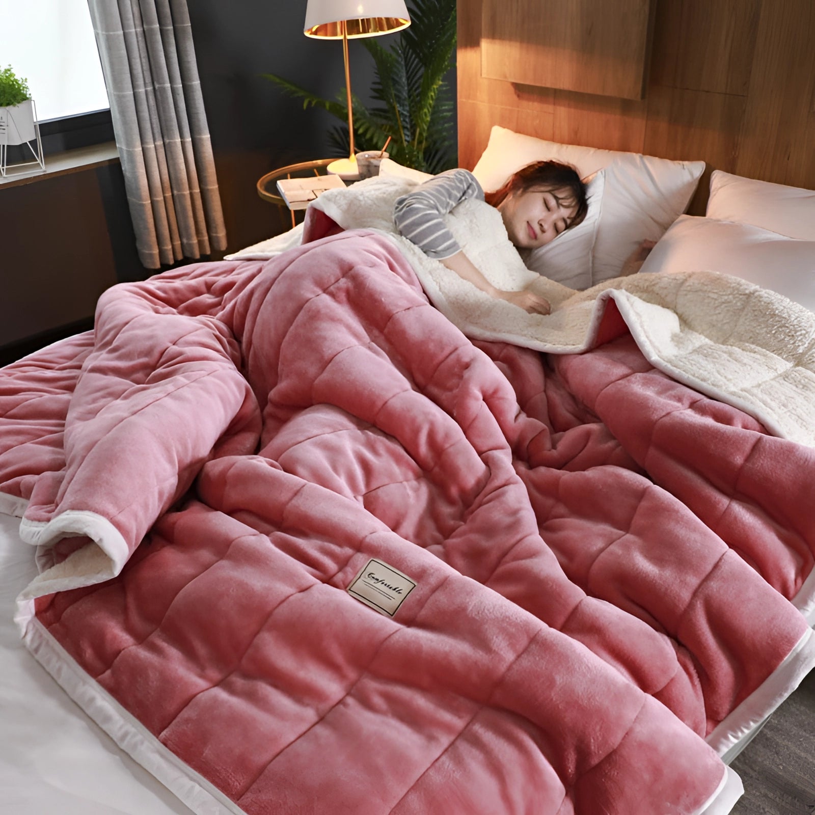 Pink plush weighted blanket draped over a bed in a cozy bedroom setting, complemented by pillows and linens, with a houseplant and a lamp visible in the background, creating a tranquil and inviting atmosphere.