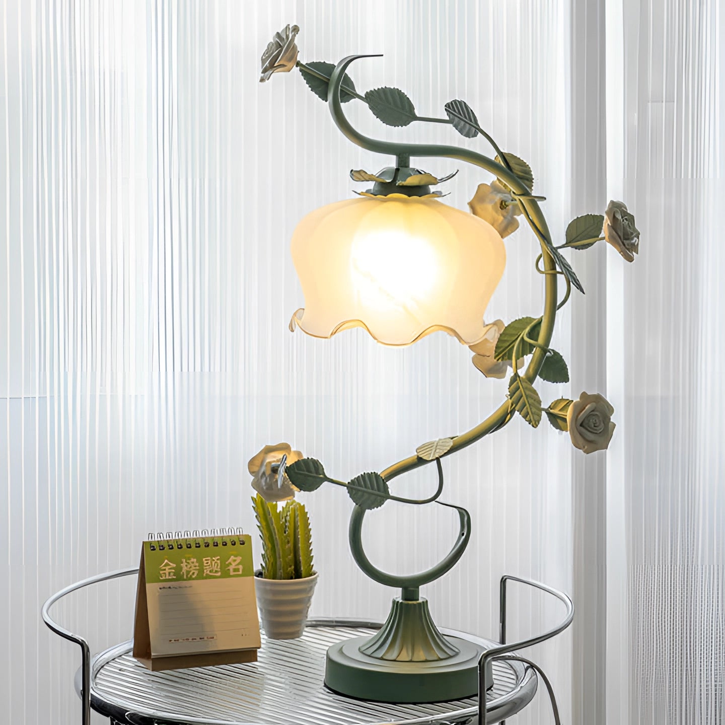 Elegant pink rose table lamp with a floral vine design, featuring green and yellow accents, placed on a table against a wall.