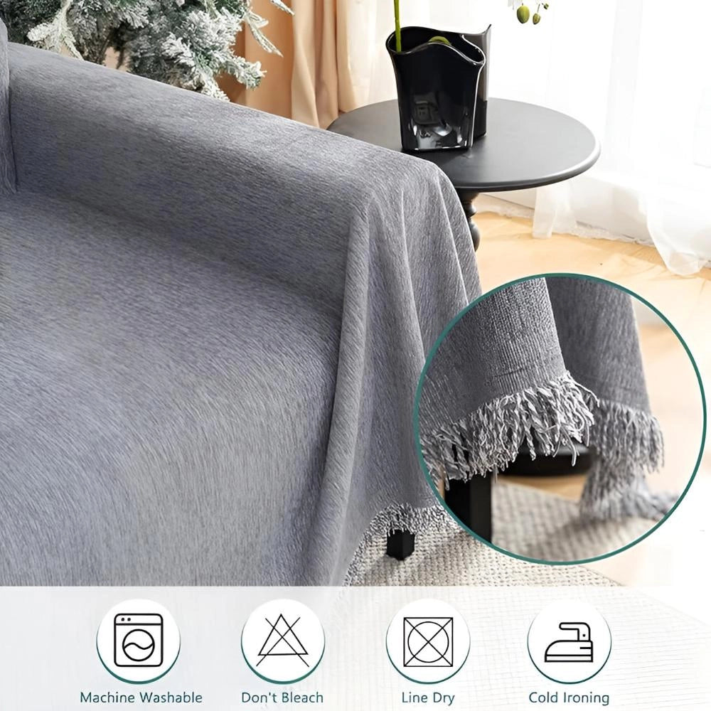 Fringed sofa blanket in grey with tassel edges draped over a sofa, showcasing its durable, comfortable texture. A plant is visible in the background, adding to the cozy ambiance.