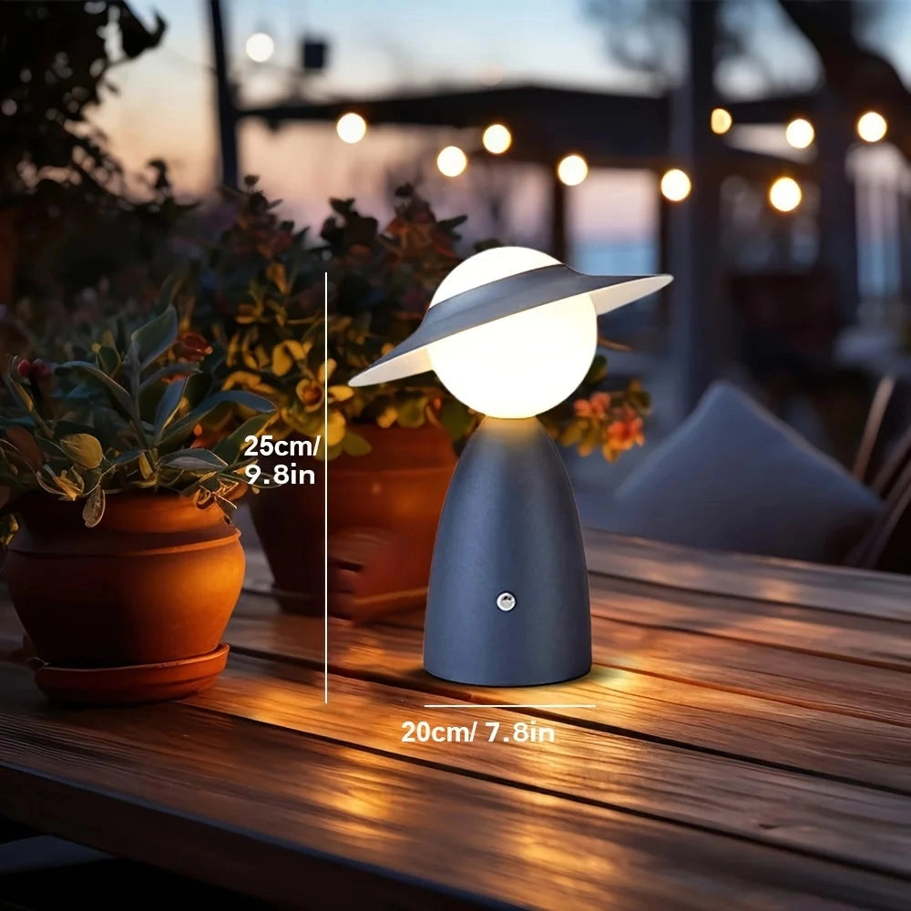 A modern gold satellite table lamp with a unique design, featuring a blue variant, sits on a wooden surface next to a potted houseplant, with a window in the background allowing natural light to filter through.