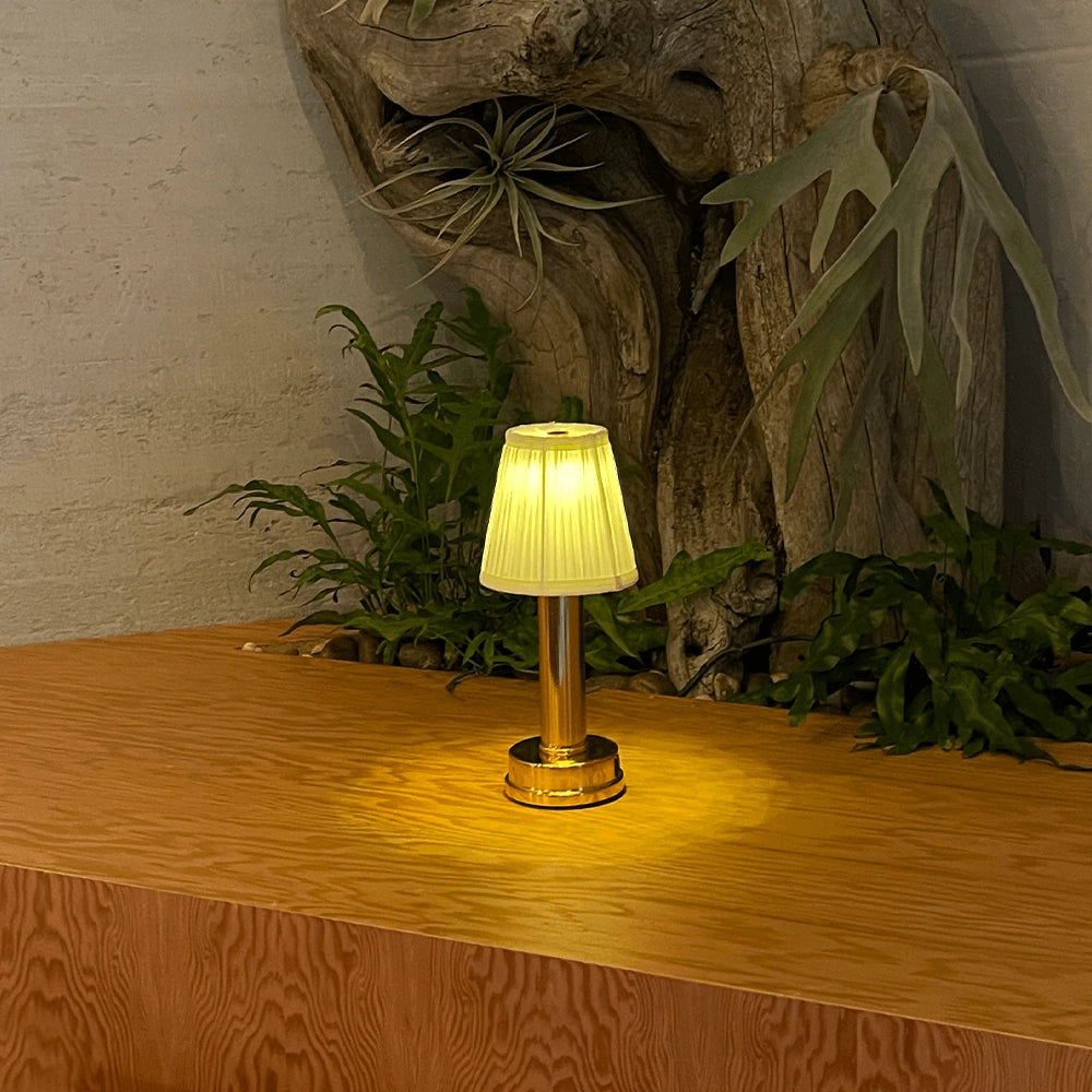 Elegant luxury designer table lamp with a gold and brass finish, placed on a wooden table. The setting includes interior design elements such as a plant and art, creating a sophisticated and stylish bedside lighting arrangement.