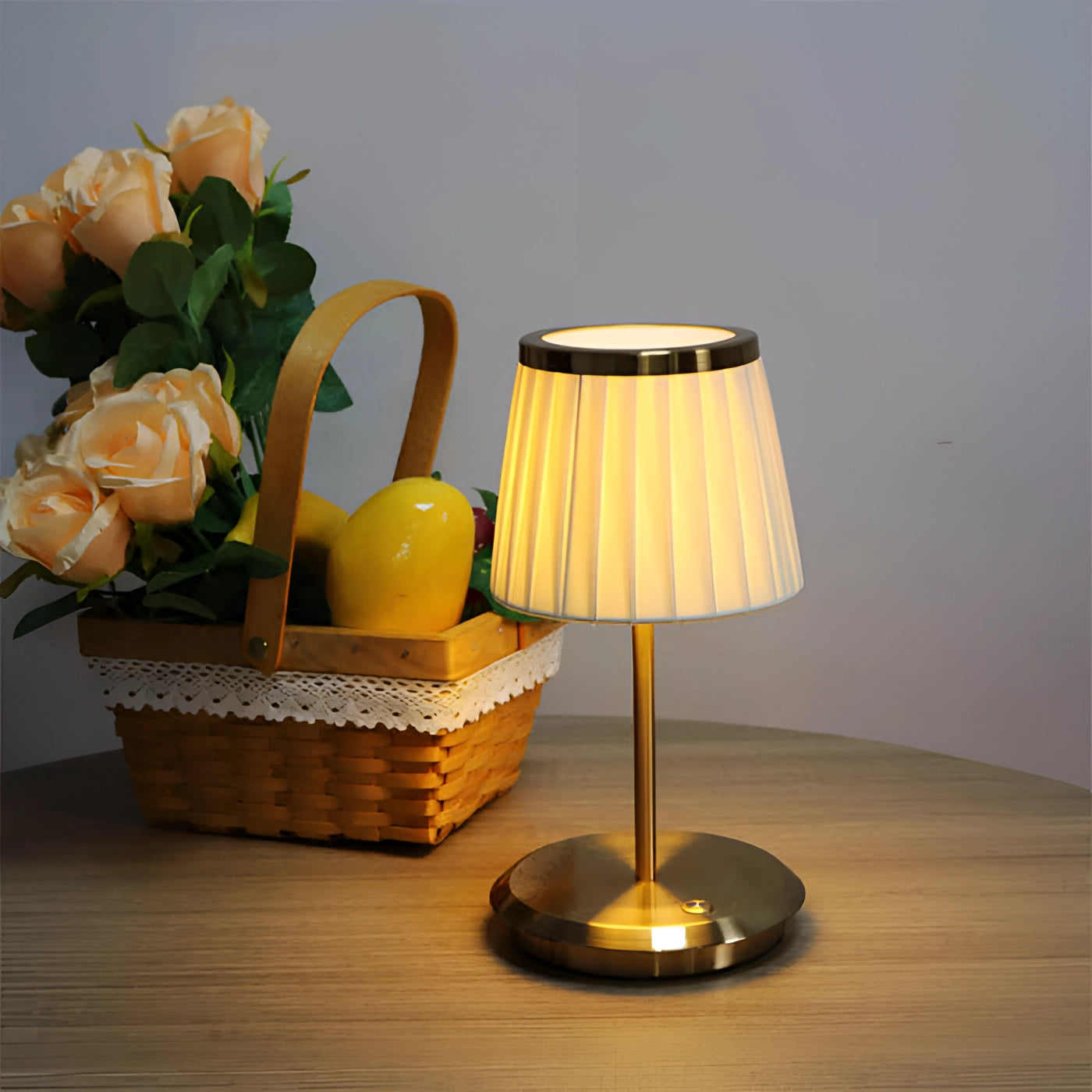 A gold table lamp with a pleated shade placed on a wooden table, surrounded by decorative elements including a potted plant, a small flower vase, and a few pieces of drinkware and tableware.