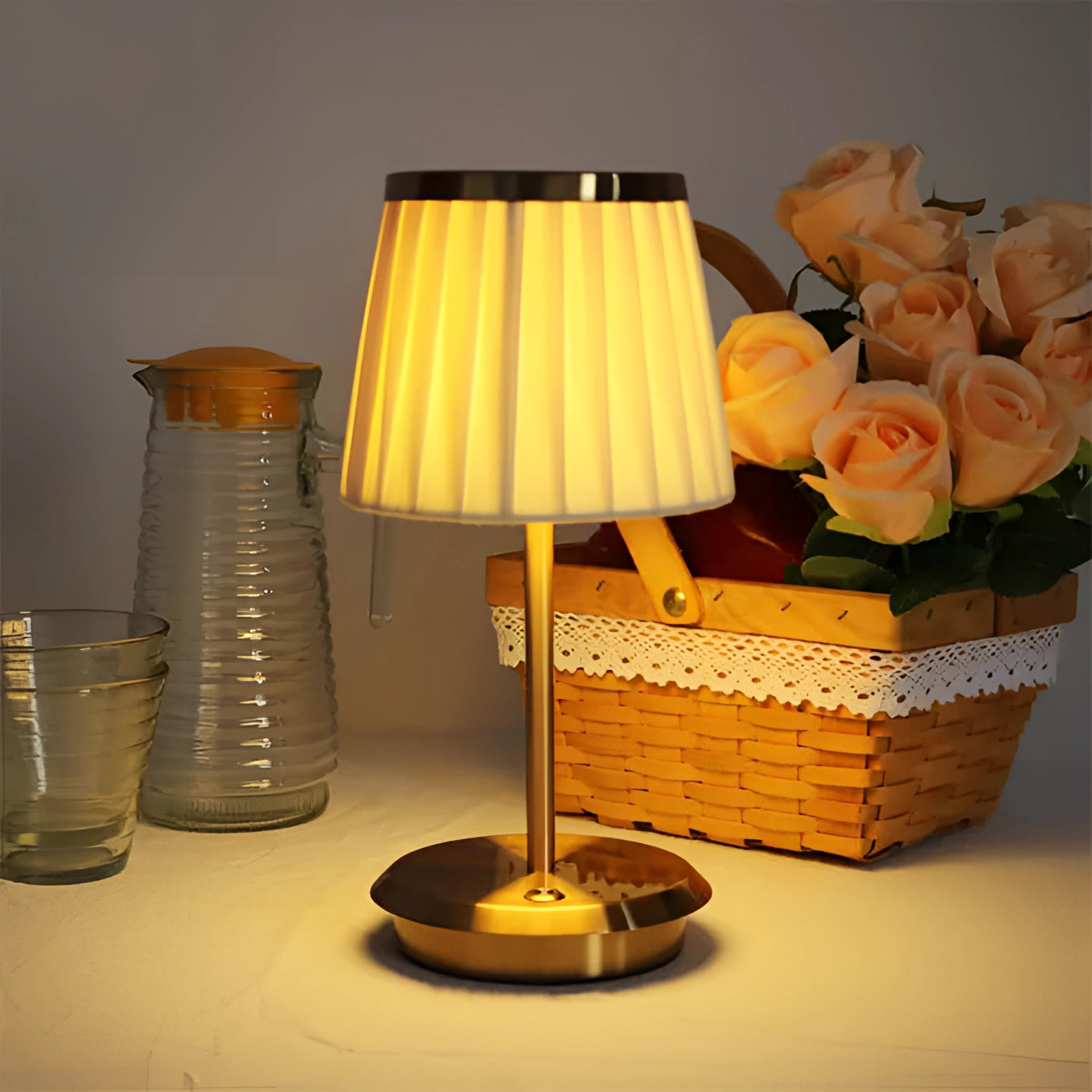 Elegant gold table lamp with pleated shade placed on a wooden table, surrounded by a decorative arrangement of roses and greenery.