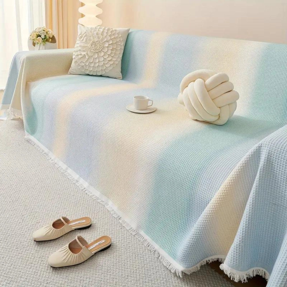 Lightweight pastel blue bedspread made of textured cotton with fringed edges, draped over a rectangular bed with white and beige pillows in a cozy bedroom setting featuring wood accents.