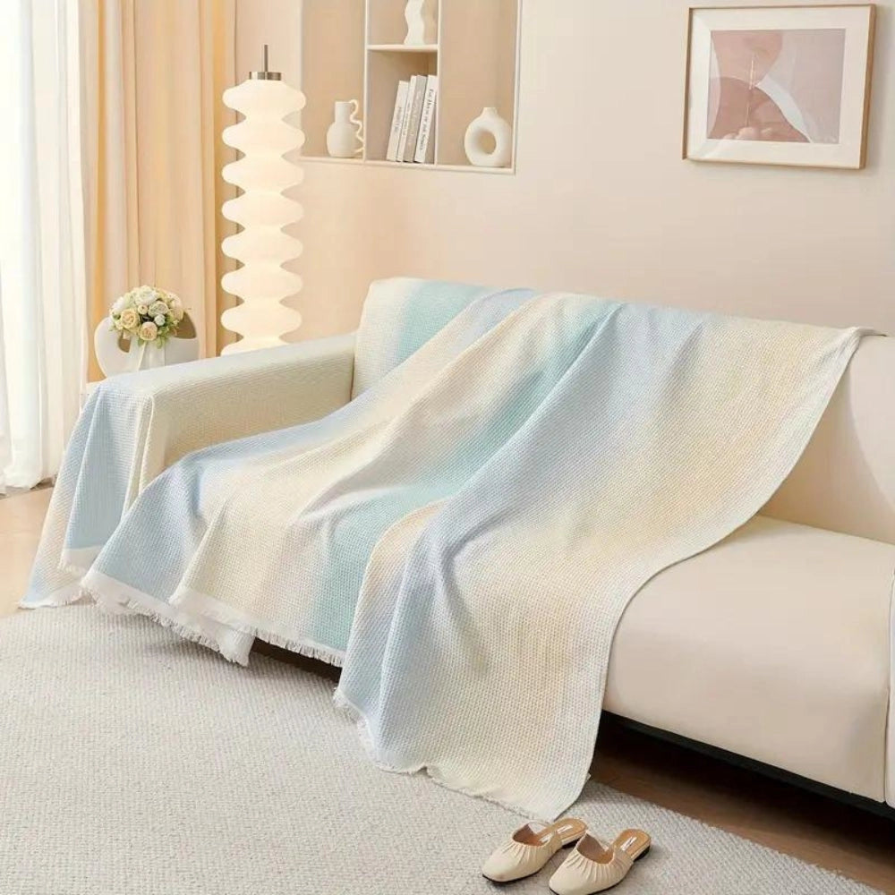 Lightweight pastel blue bedspread made of textured cotton with fringed edges, draped over a couch in a cozy interior setting.