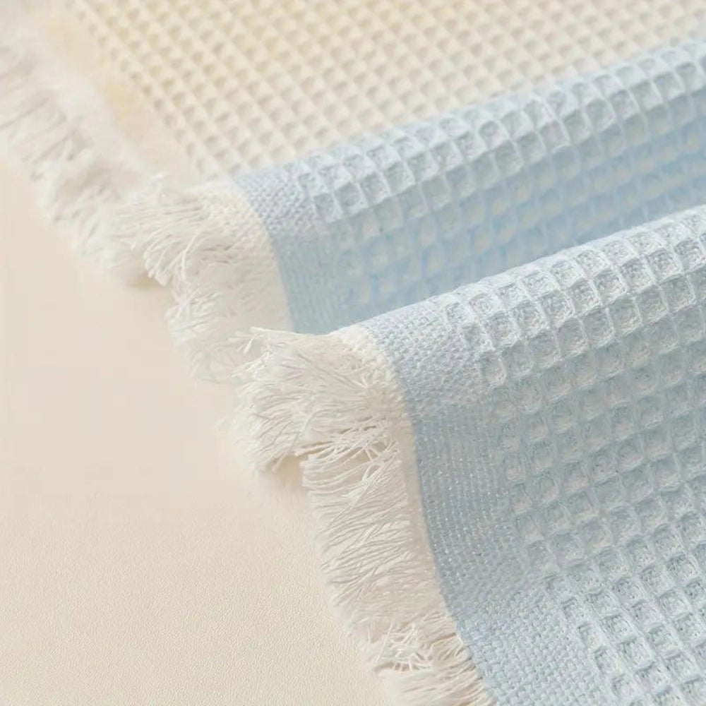 Lightweight pastel blue bedspread made from textured cotton featuring fringed edges, draped over a wooden floor.