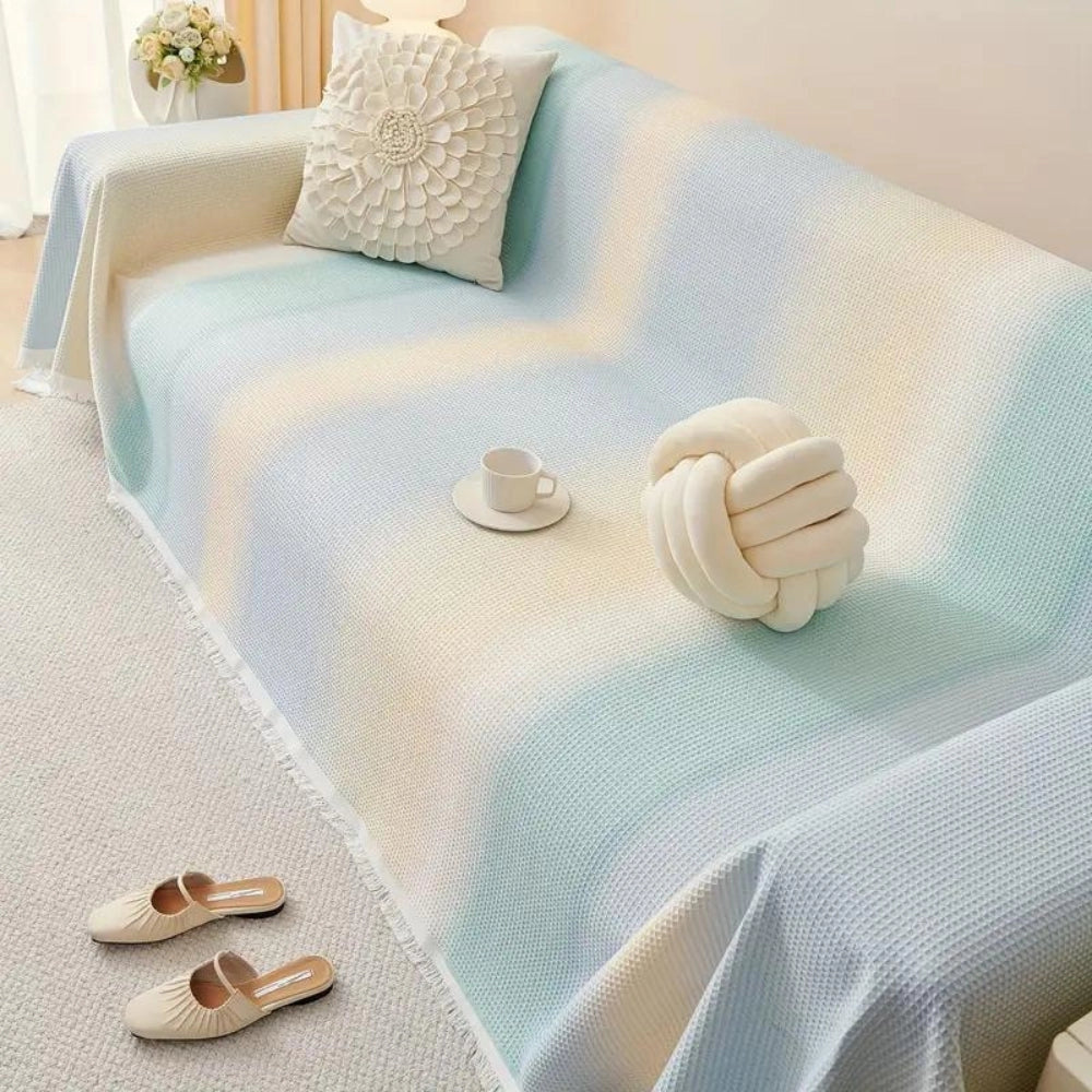 Lightweight pastel blue textured cotton bedspread with fringed edges draped over a four-seater setup (180x300cm), contrasting against a beige and wooden interior design, offering a sense of comfort and style.