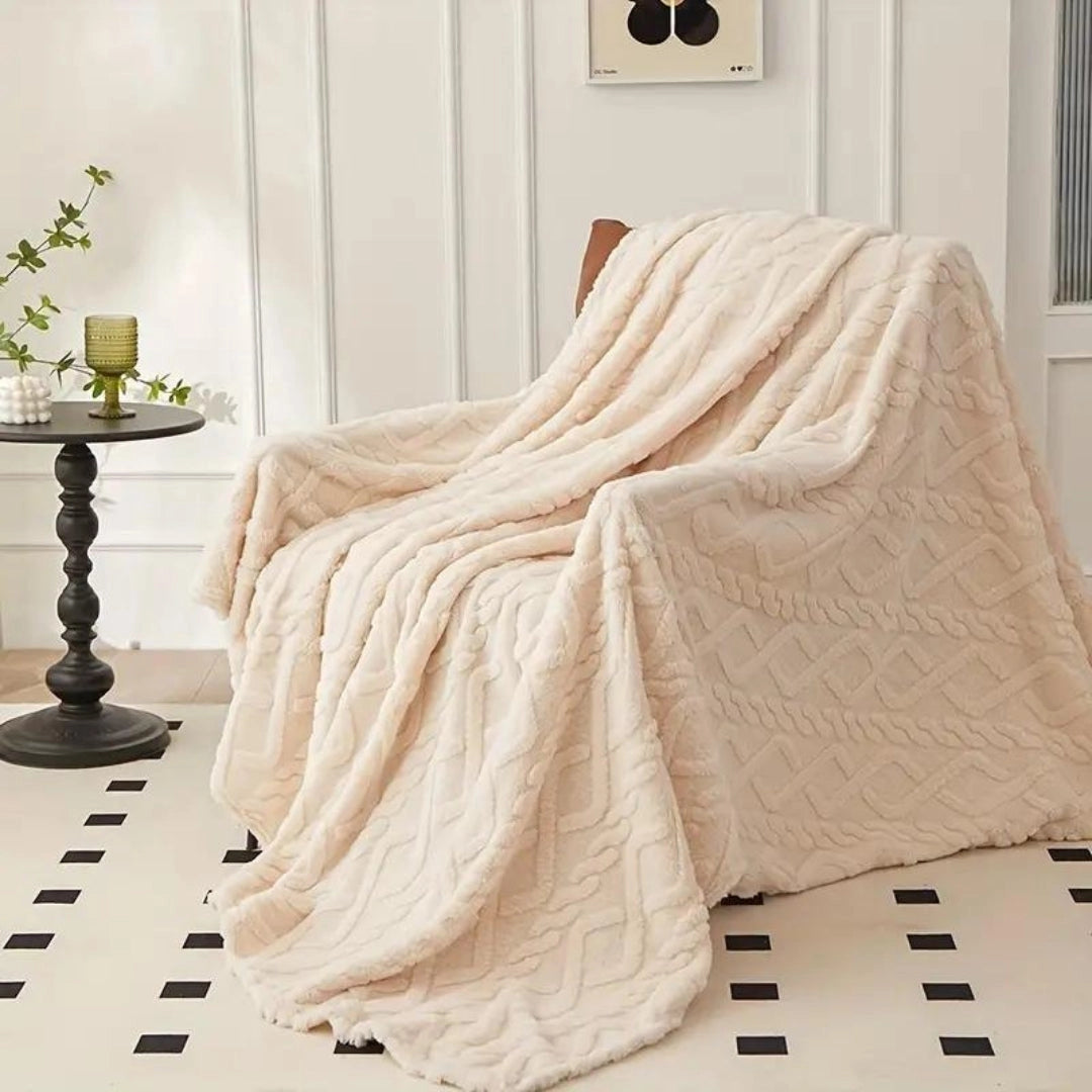 Elegant cream luxury cable knit throw blanket draped over a wooden floor next to a beige flowerpot with a houseplant, showcasing a cozy and high-quality design.