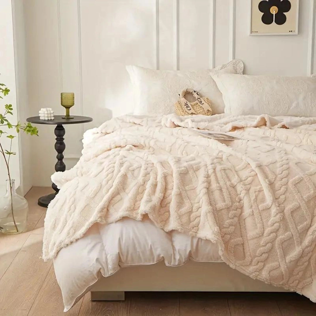 Elegant cream luxury cable knit throw draped over a bed with a wooden frame, surrounded by cozy pillows and a decorative plant in a stylish interior setting.