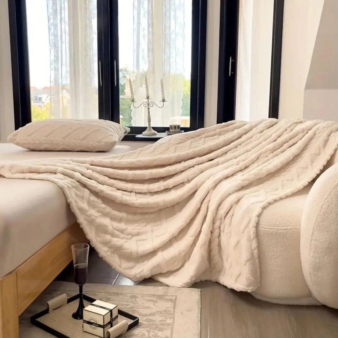 Cream-colored luxury cable knit throw draped over a piece of furniture in a cozy, elegantly designed interior space with wooden flooring and natural light streaming through a nearby window.