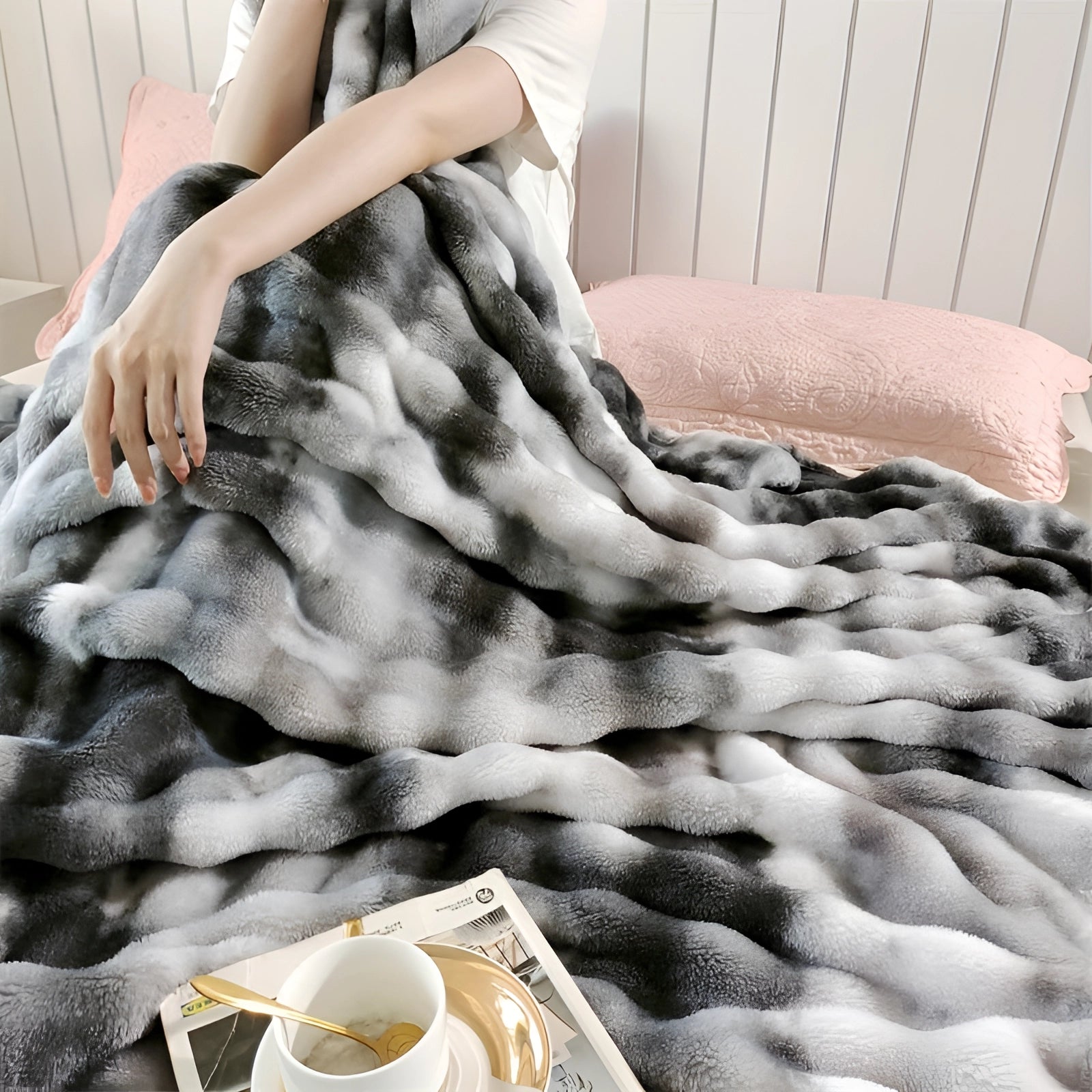 Cozy scene featuring a luxurious faux fur blanket in a plush setting; a person's arm is resting on the blanket, holding a cup, suggesting comfort and relaxation.