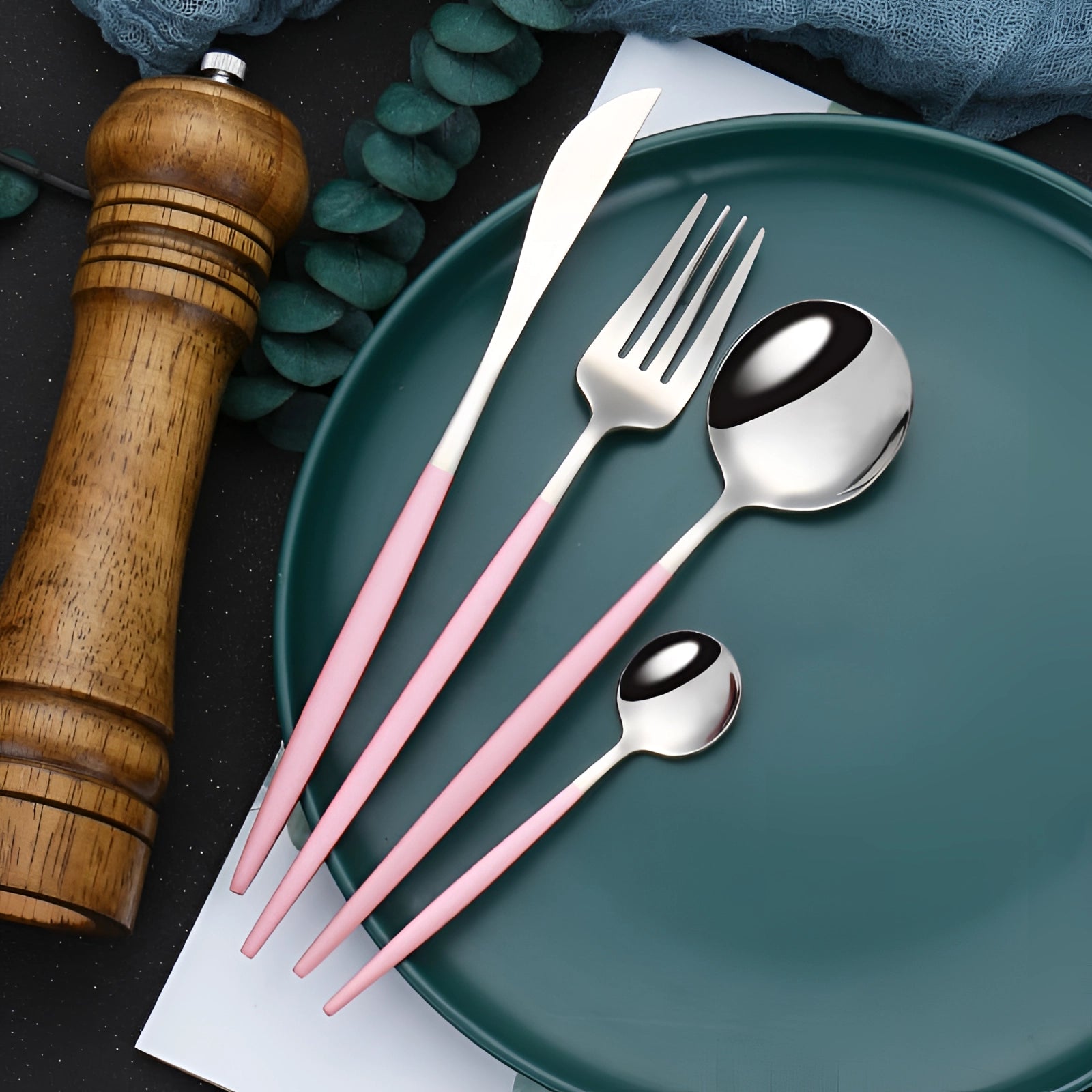 Elegant luxury cutlery set in gold with modern design, featuring a table knife and other utensils displayed alongside a pink and silver theme, set on a wooden table with a circular plate.