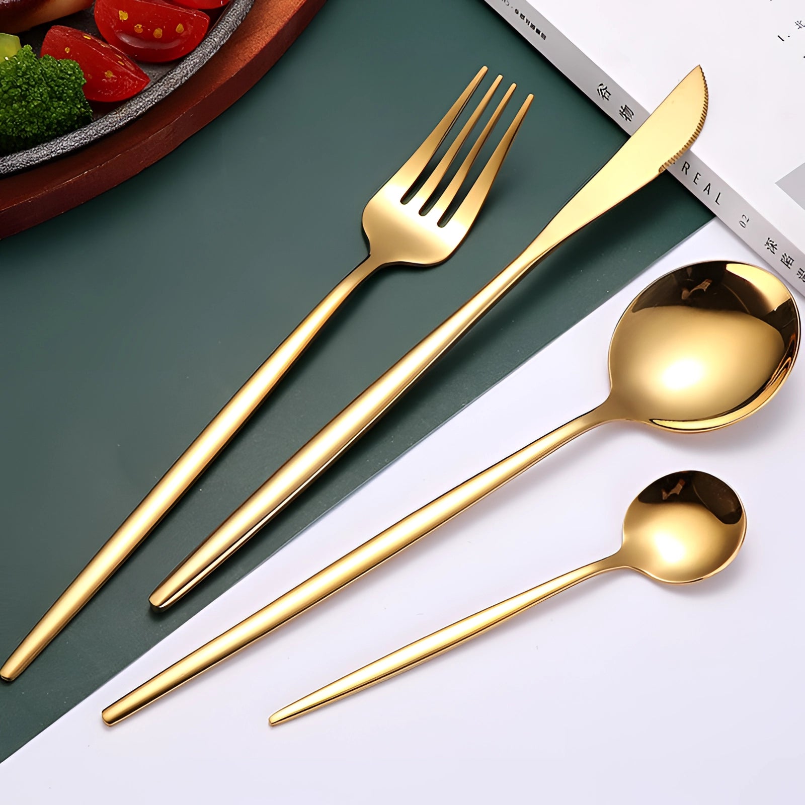 Elegant luxury gold cutlery set displayed on a wooden table, featuring modern design elements with forks, knives, and spoons complementing the tableware. The setting includes artistic arrangements of dishes and leaf vegetables, highlighting the sophisticated ambiance suitable for fine dining.