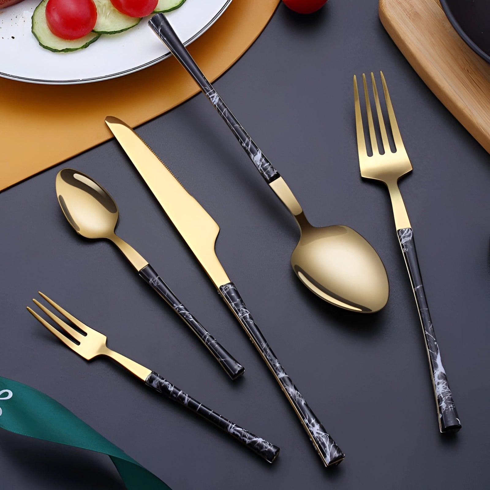 Luxury gold marble handle cutlery set consisting of 5 pieces, including a fork, displayed elegantly on a dining table setting with a plate and plum tomato.