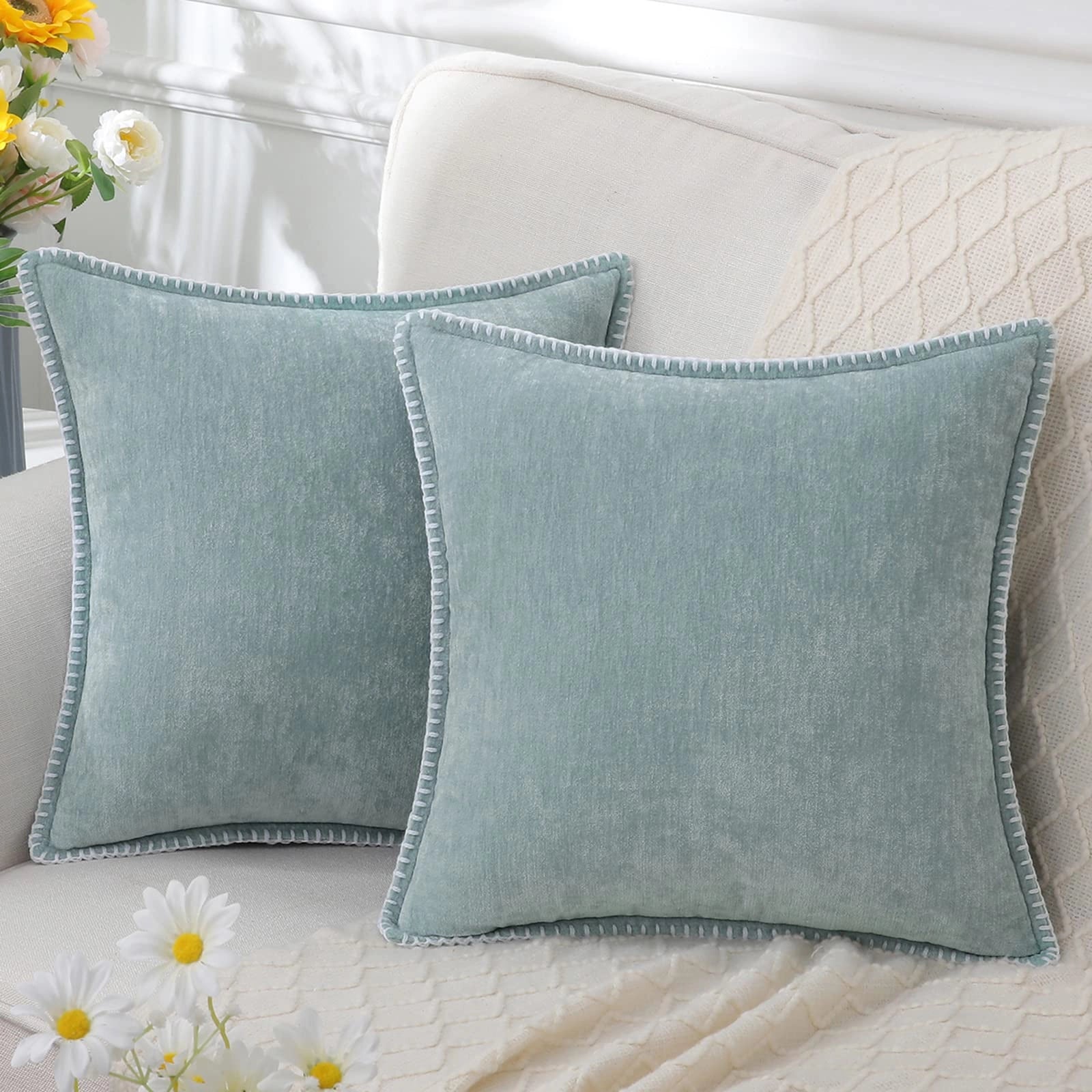 Light blue luxury velvet throw pillows arranged on a grey couch with a plant and floral decor in the background.