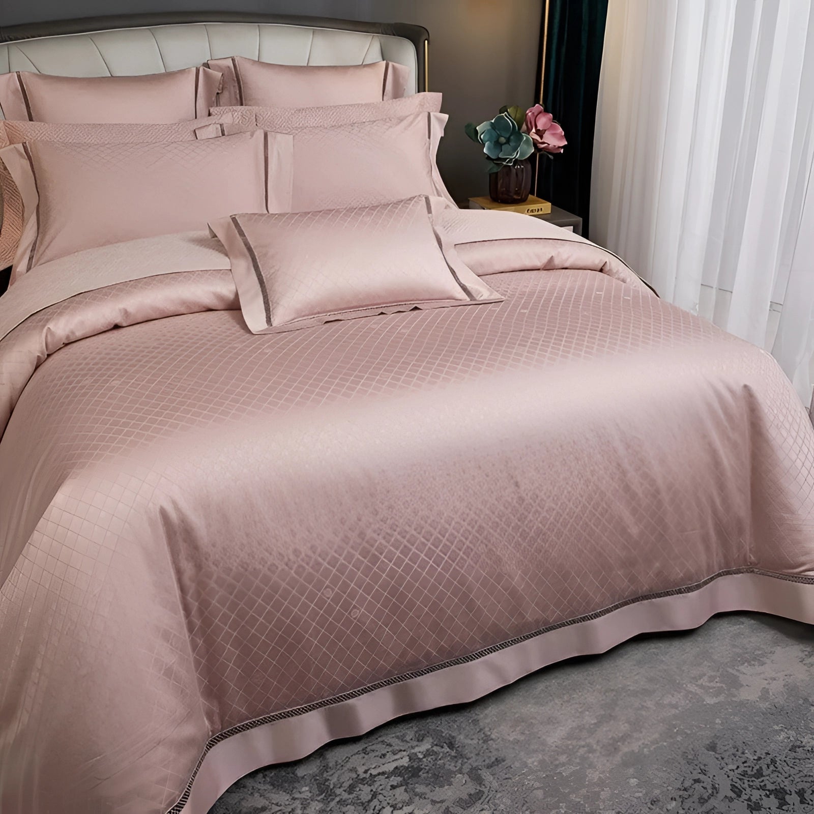 Luxurious pink satin bedding set with a geometric design, including a duvet cover and matching pillowcases, displayed on a wooden bed frame with hardwood flooring, accompanied by a bedside lamp and a small green plant.