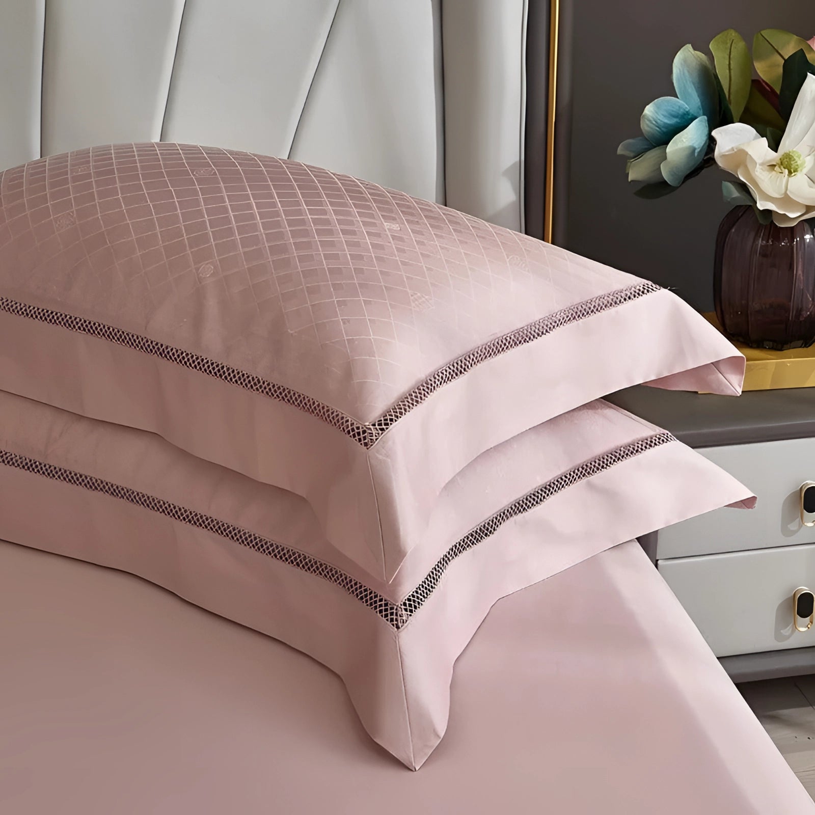 Luxury pink satin bedding set featuring a geometric pattern, with a duvet and matching pillowcases adorned on a neatly arranged bed. The setting includes surrounding wooden furniture, such as a chest of drawers, complementing the elegant and comfortable bedroom decor.