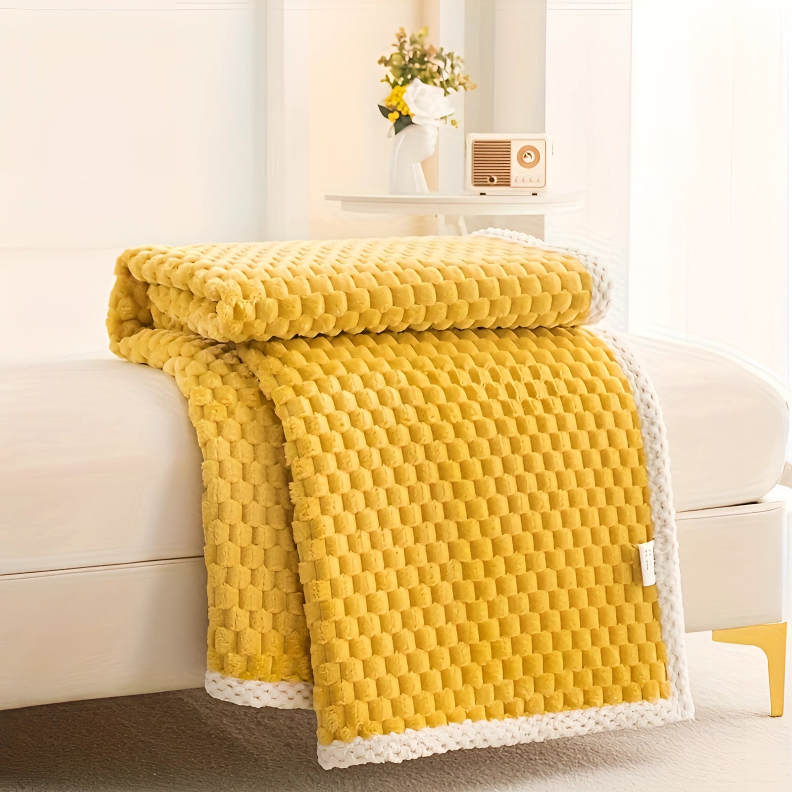 Luxury pink textured blanket draped over a chair in a cozy, modern interior setting, featuring a plush knitted design with a pom-pom trim, showcased in a variant color of yellow, measuring 200 x 230cm, surrounded by furniture including a wooden table and a plant, emphasizing comfort and style.