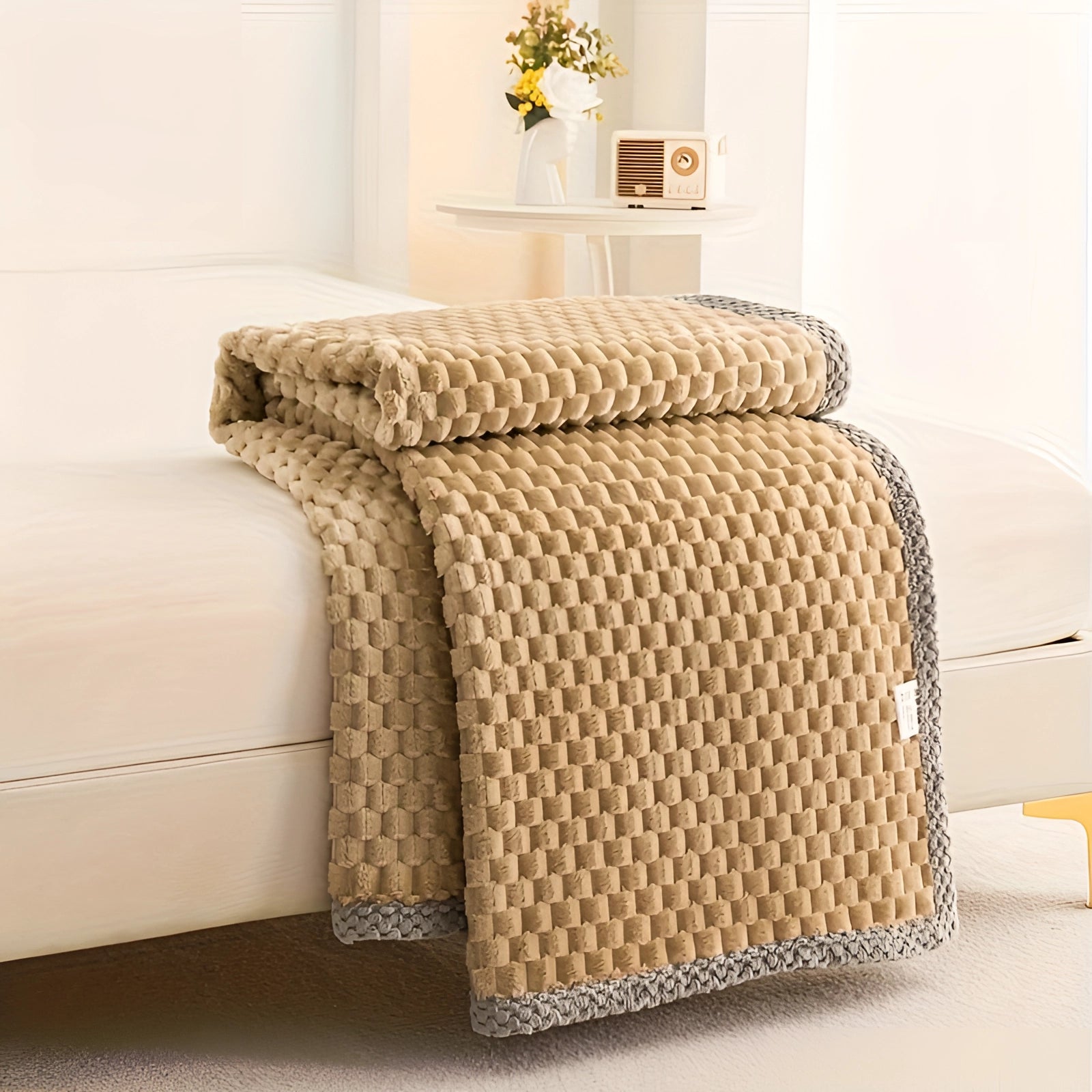 Luxury camel-colored textured blanket with pom-pom trim draped over a beige bed, set in a cozy interior with wooden flooring and a plant in the background.