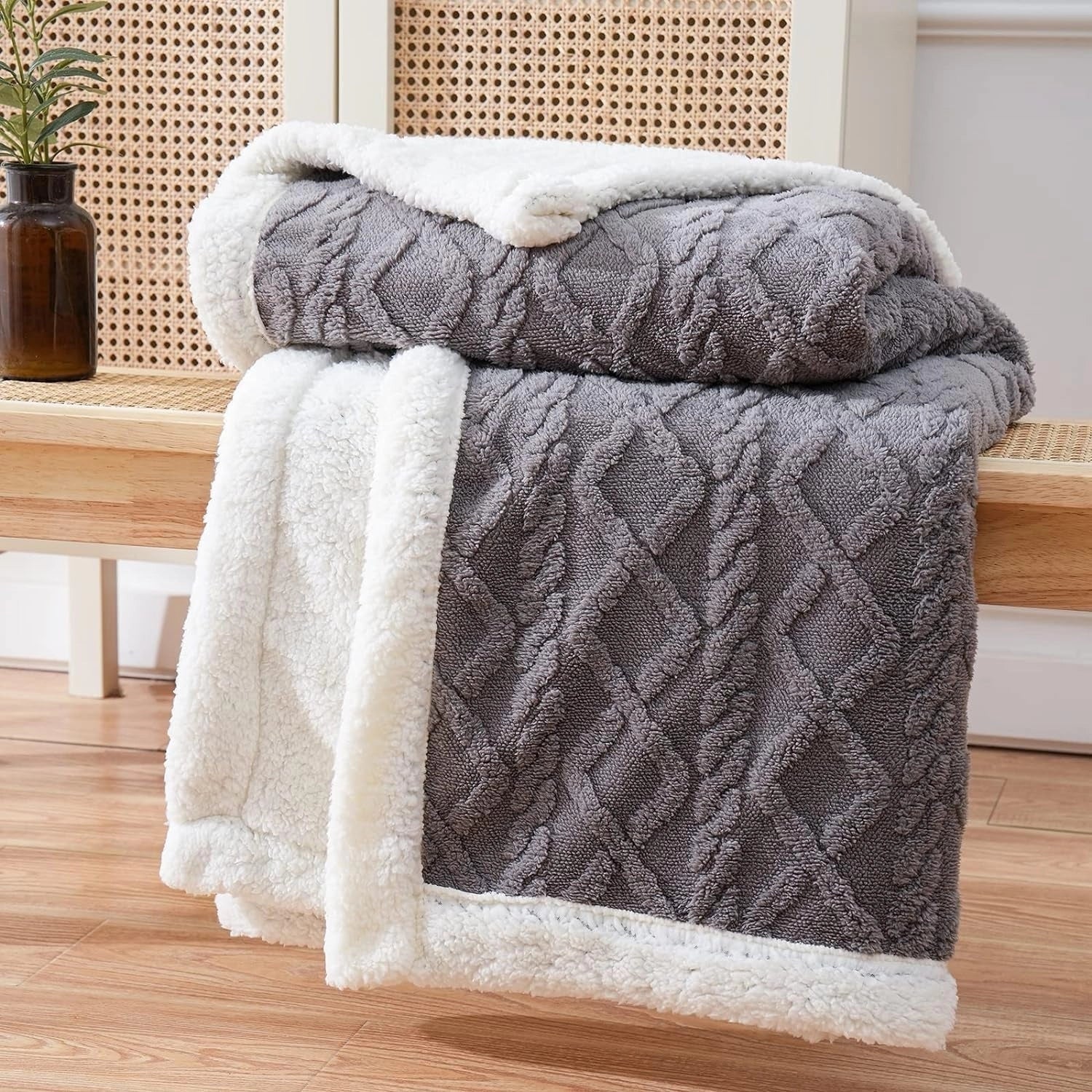 A luxurious gray reversible Sherpa fleece blanket draped over a hardwood floor near a window, with a cozy ambiance enhanced by a nearby flowerpot and houseplant.