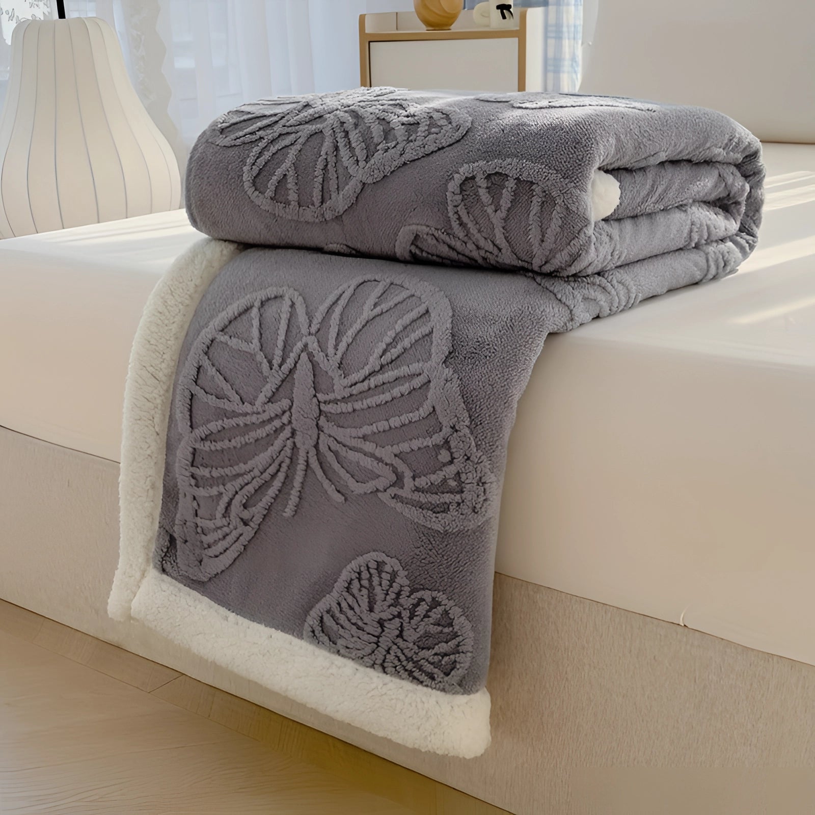 Luxury Sherpa Fleece Blanket in plush gray with leaf pattern, draped over a bed in a dark gray color featuring a cozy and comfortable textile. The scene includes a pillow and wooden elements, creating a warm and inviting interior design atmosphere.