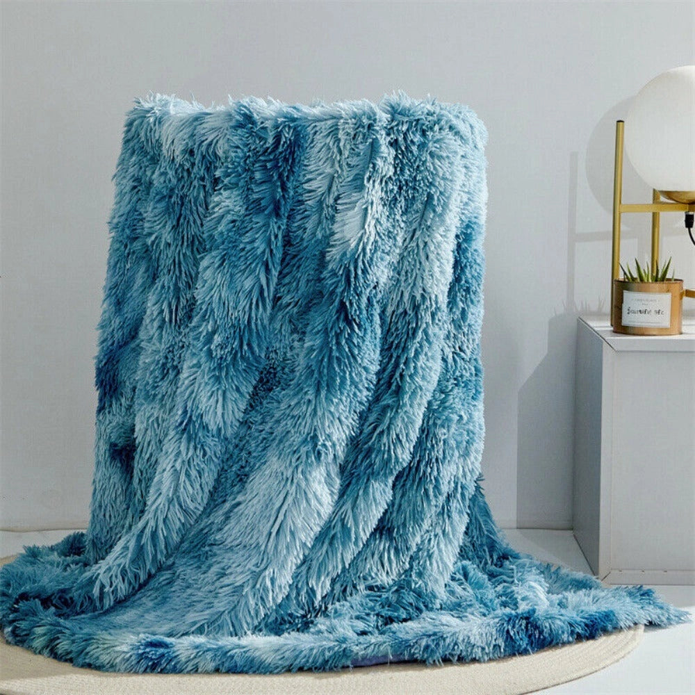 Luxurious white faux fur blanket with a plush and textured design, elegantly draped over a surface, displaying a hint of ocean blue. The large rectangular blanket measures 200x300cm and is set against a backdrop featuring wooden elements, grey tones, and decorative plants, creating a cozy and sophisticated ambiance.