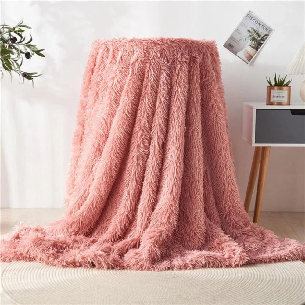 Luxury rose pink faux fur blanket with a textured plush fabric, elegantly draped over a wooden floor, surrounded by houseplants, showcasing a cozy and stylish setting.