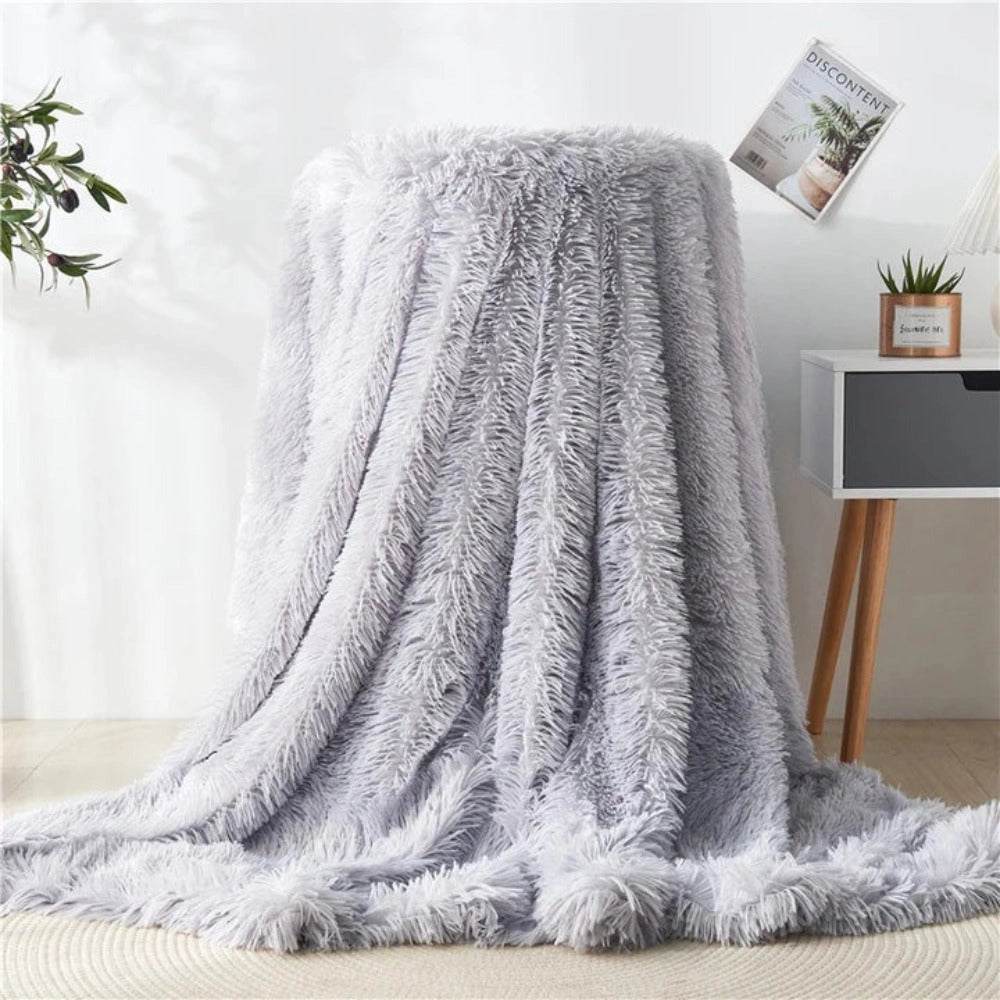 Light grey luxury faux fur blanket draped elegantly over a wooden surface, with a textured plush design, creating a cozy and sophisticated atmosphere. A window with plants and trees outside can be seen in the background, adding a touch of nature to the scene.