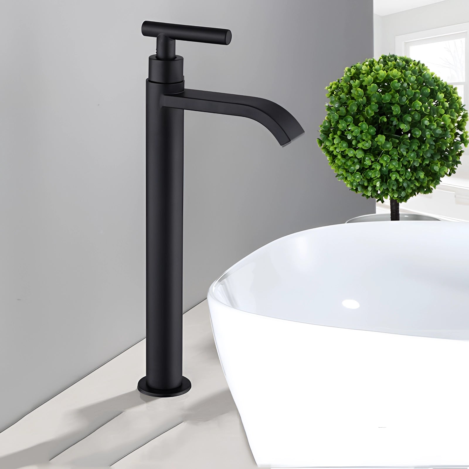 Matte black modern bathroom faucet with a sleek minimalist design, installed on a rectangular sink, surrounded by potted houseplants on a light-colored table.