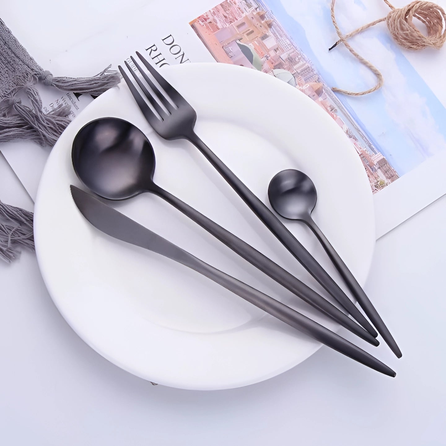 A sleek matte black stainless steel cutlery set displayed neatly on an elegant table setting with a white plate, featuring a table knife, fork, and spoon, arranged on a soft linen tablecloth.