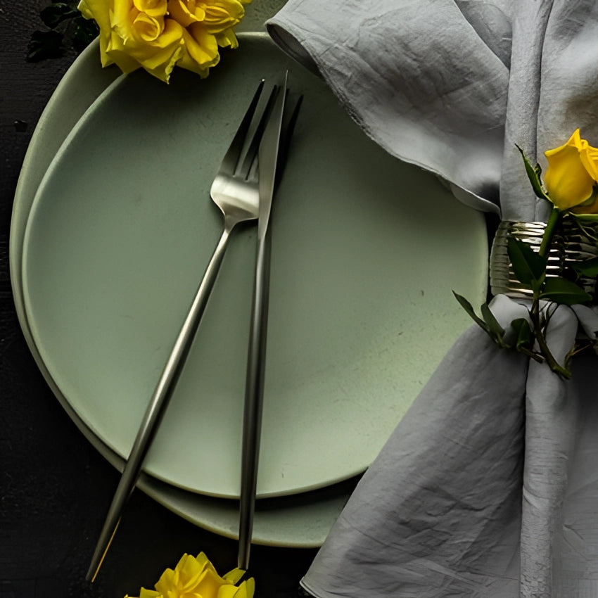 Matte black stainless steel cutlery set on a modern minimalist table setting with yellow flowers and petals, grey textile tablecloth, and matching dishware.