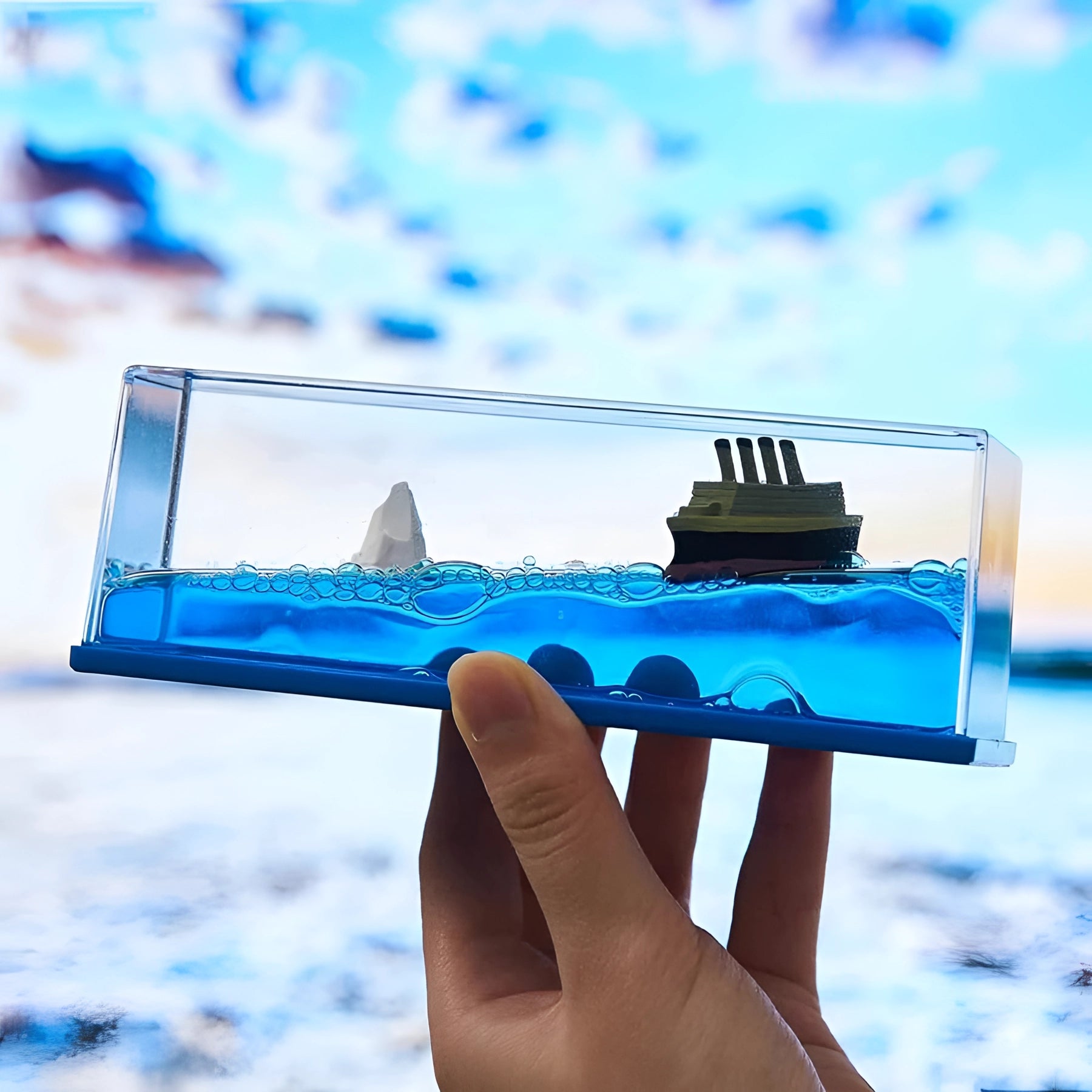 
A hand holding a miniature ship encased in a clear acrylic wave, against a backdrop of a blue sky with clouds.