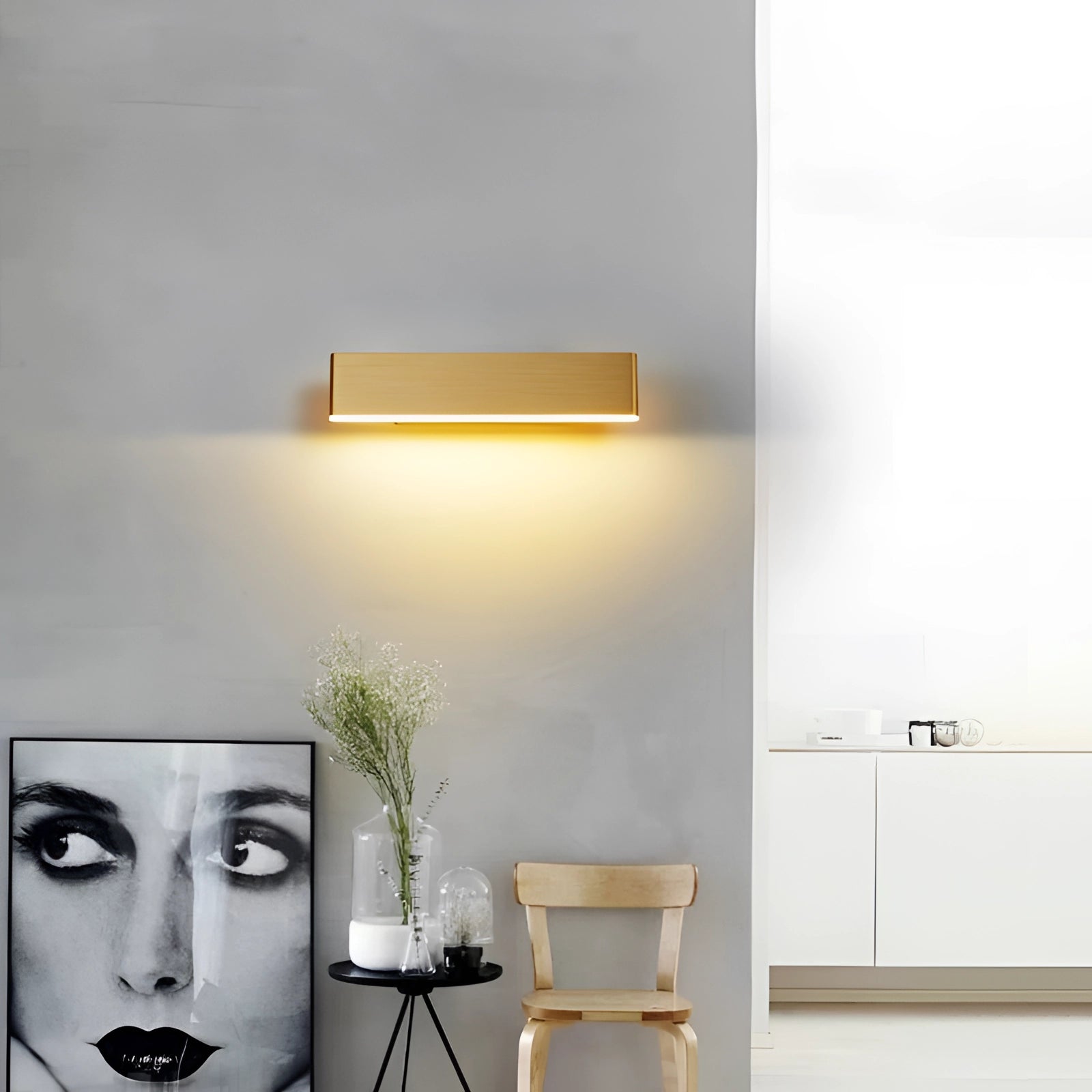 Modern black and gold rectangular LED wall sconce with a minimalist geometric design, emitting cold white light, mounted on a grey wall next to a wooden shelf with a small plant.