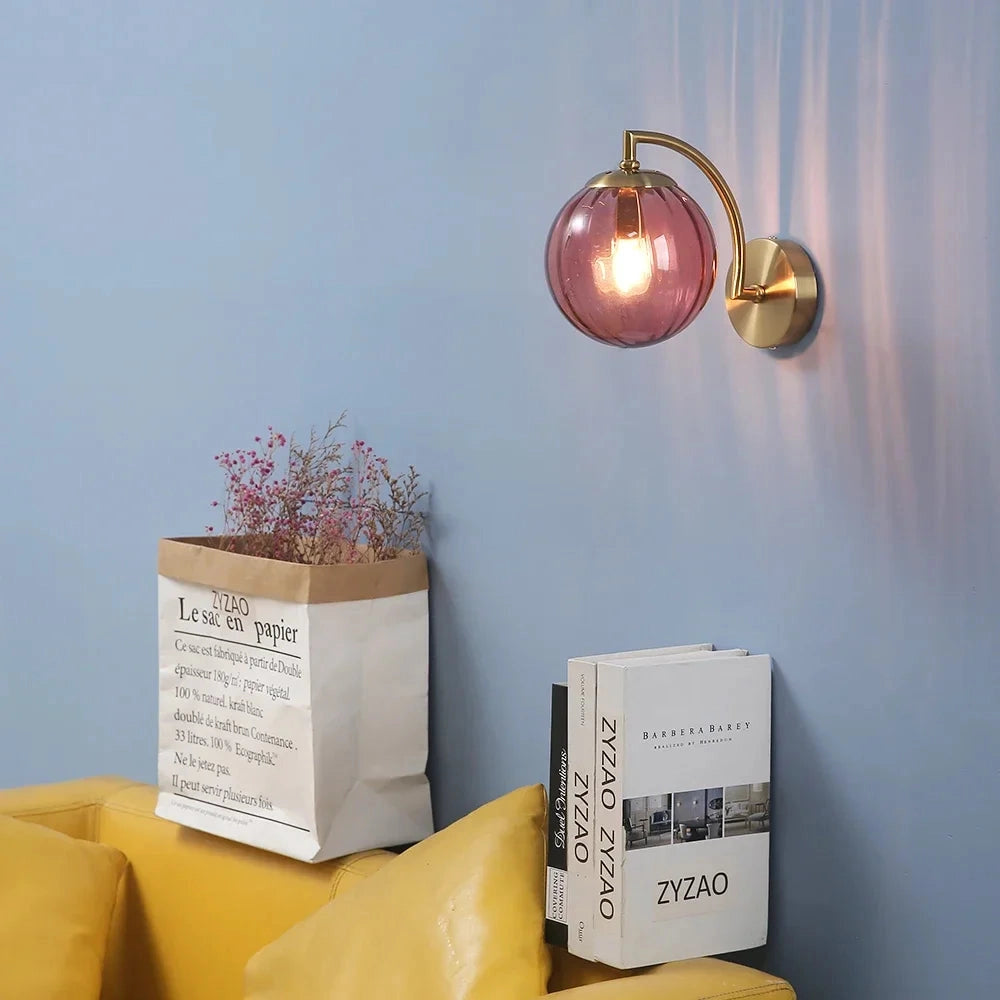 Modern brass globe wall sconce with a 22.7cm wide decorative lamp mounted on a wooden panel, illuminating a room with warm yellow lighting, set against an interior design backdrop with plants.