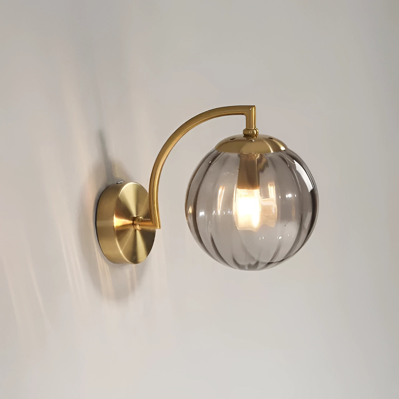 Modern brass globe wall sconce with a smoke grey finish, featuring a decorative circular design. The sconce includes a light bulb encased within an amber-tinted globe, mounted on a metal base.