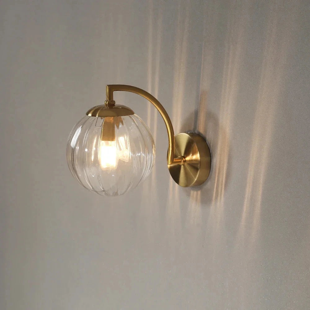 Modern brass globe wall sconce with a 22.7cm wide decorative lamp, featuring a spherical design with a smooth metal finish. The lamp is mounted on a wooden wall, with a soft warm glow from an exposed light bulb, surrounded by subtle plant decorations.