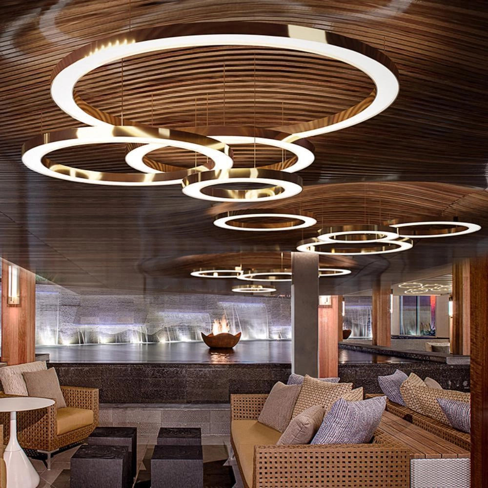 Circular LED ring pendant light hanging from the ceiling in a luxury hotel lobby, with a modern interior design featuring a couch and wooden architectural elements.