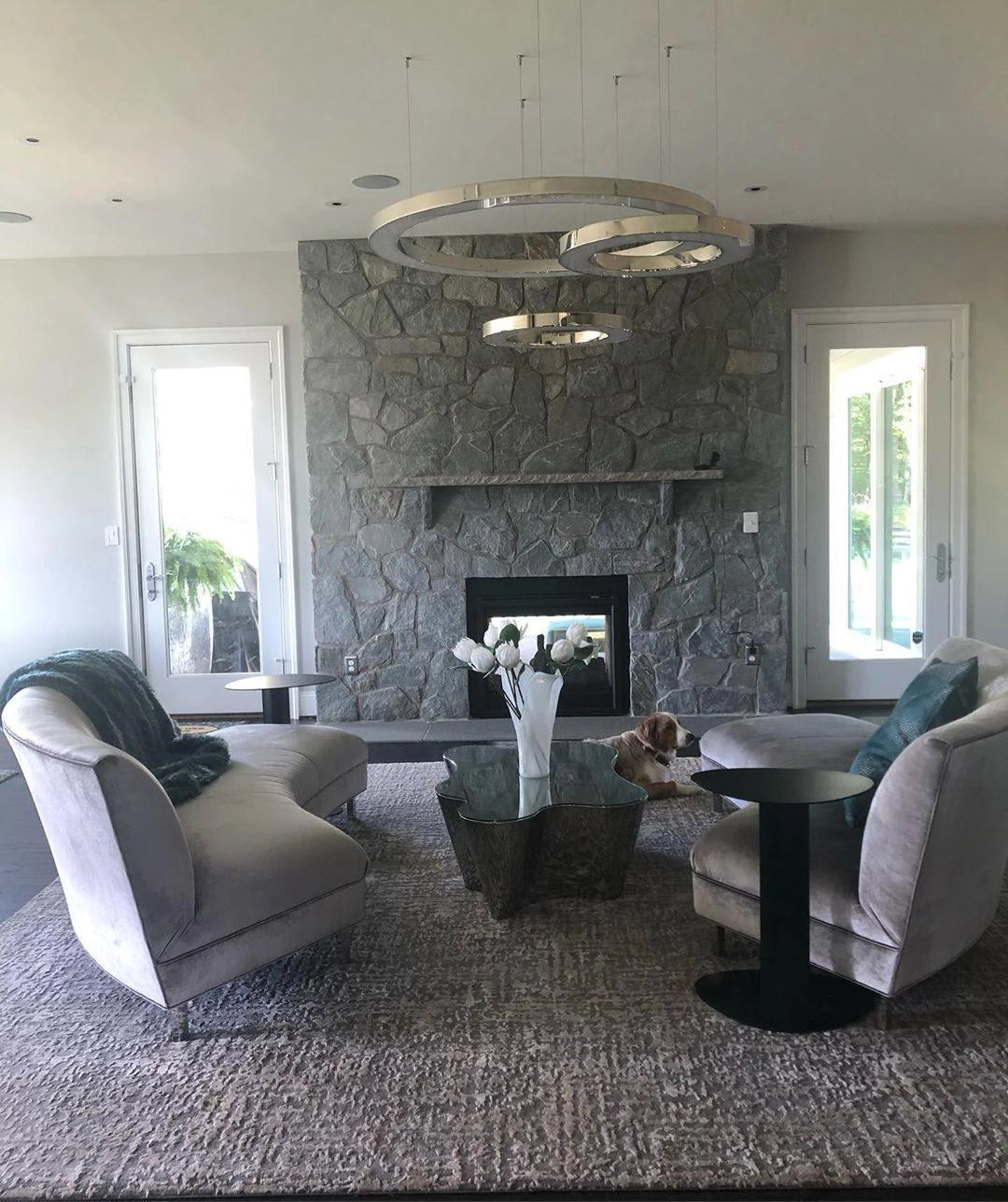 Modern circular LED ring pendant light hanging from the ceiling in a luxurious hotel lobby setting, with surrounding furniture including chairs, tables, and couches, large windows allowing natural light, and wooden accents in the room.