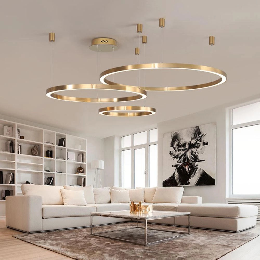 A modern circular LED ring pendant light hangs from the ceiling, illuminating a luxurious hotel lobby setting with contemporary furniture including a white couch and wooden table, surrounded by elegant interior design elements and flooring.