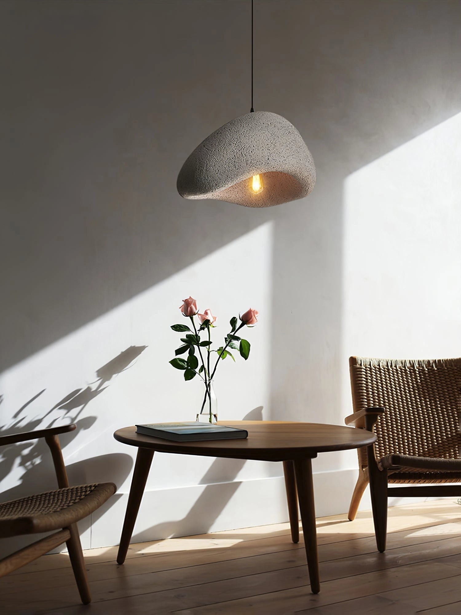 Modern gray curved pendant light with a minimalist sculptural design, hanging elegantly in a well-furnished interior setting featuring a wooden table, a potted plant, and a stylish chair.