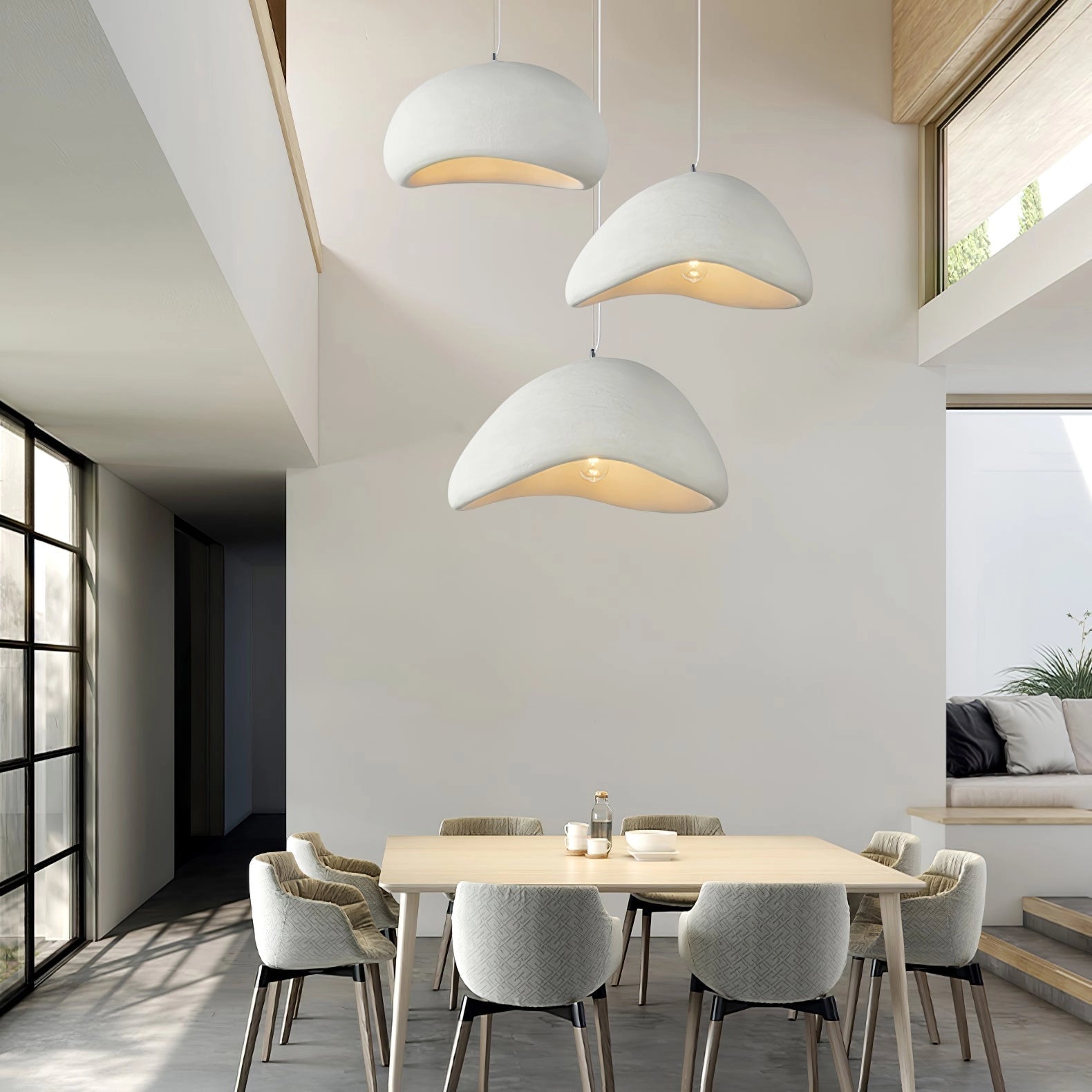 A modern curved pendant light with a minimalist sculptural design in a white finish, measuring 50cm in diameter and 30cm in height, hanging in a contemporary interior setting featuring a wooden floor, a table, chairs, and a plant.